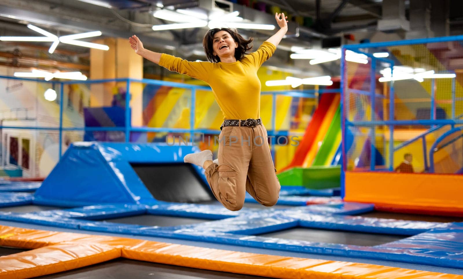 Pretty girl jumping on colorful trampoline at playground park and smiling. Beautiful young woman during active entertaiments