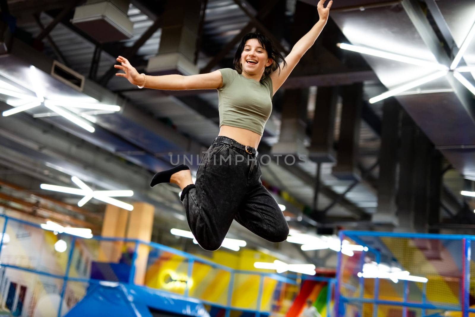 Pretty girl jumping on colorful trampoline at playground park and smiling. Beautiful female teenager happy during active entertaiments indoor