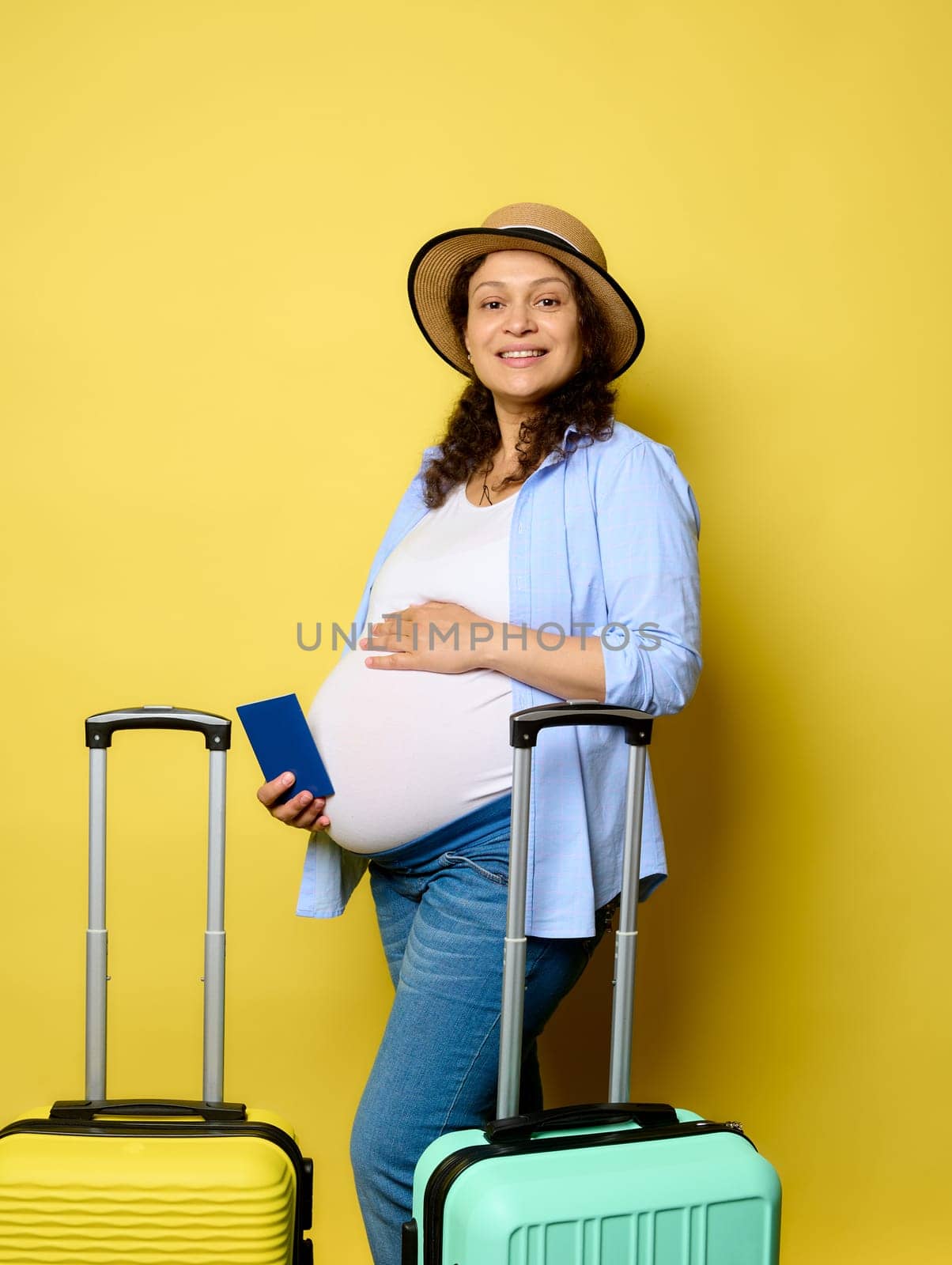 Happy curly haired multi-ethnic pregnant woman in stylish casual clothes, stroking belly, traveling abroad during pregnancy, smiling cutely looking at camera, isolated on yellow studio background