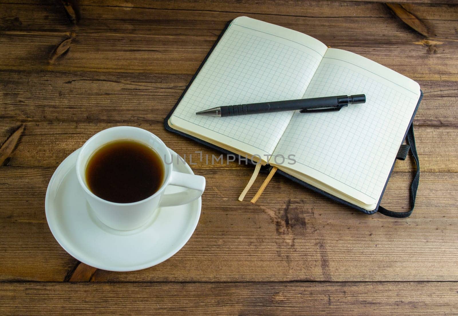 Coffee in a cup, notebook and pen. Coffee in a cup with a saucer and a notebook with a pen on a wooden table.