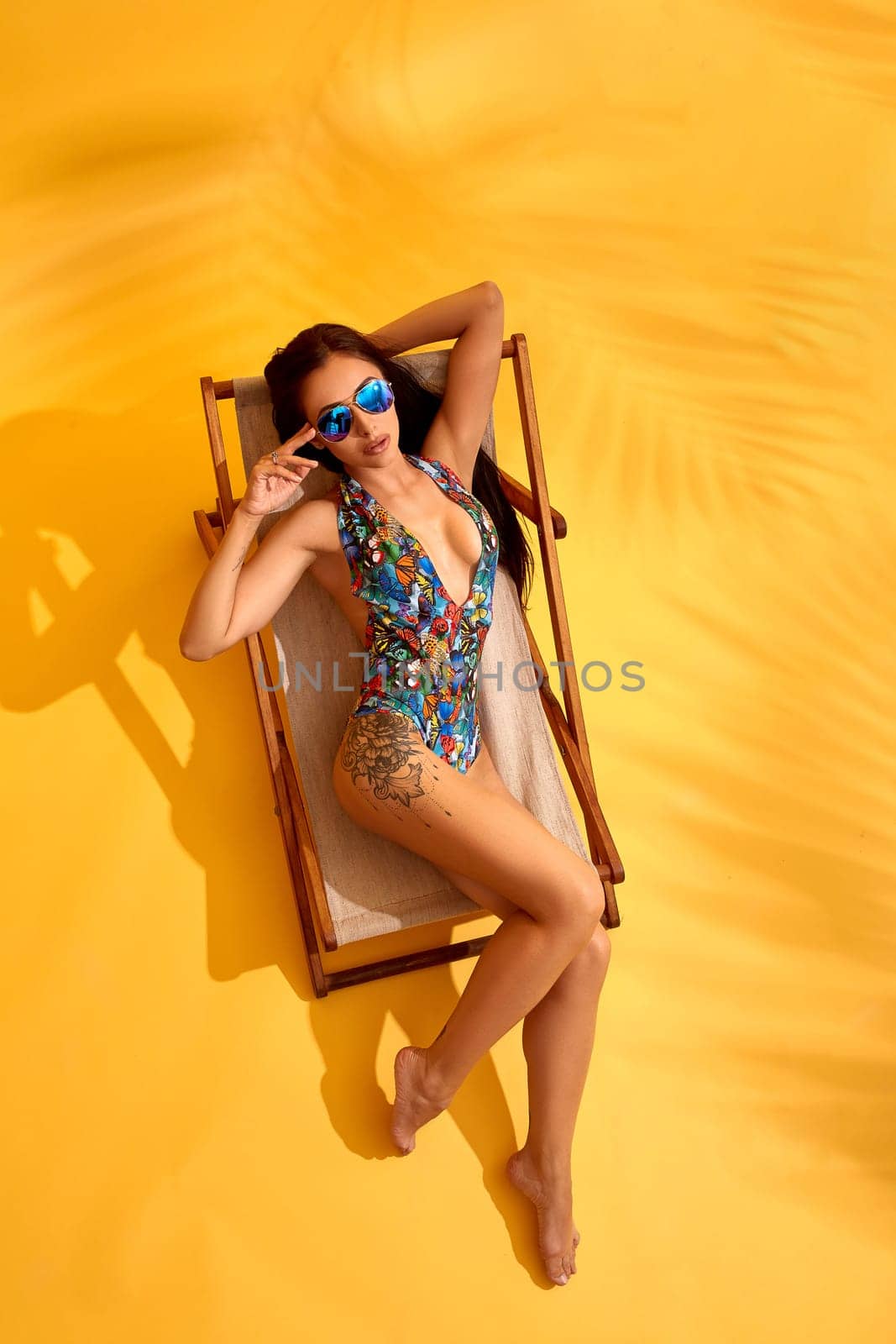 Beautiful young brunette woman in colored swimwear lies on the lounger on yellow background. Blue sunglasses, hands behind head. Top view