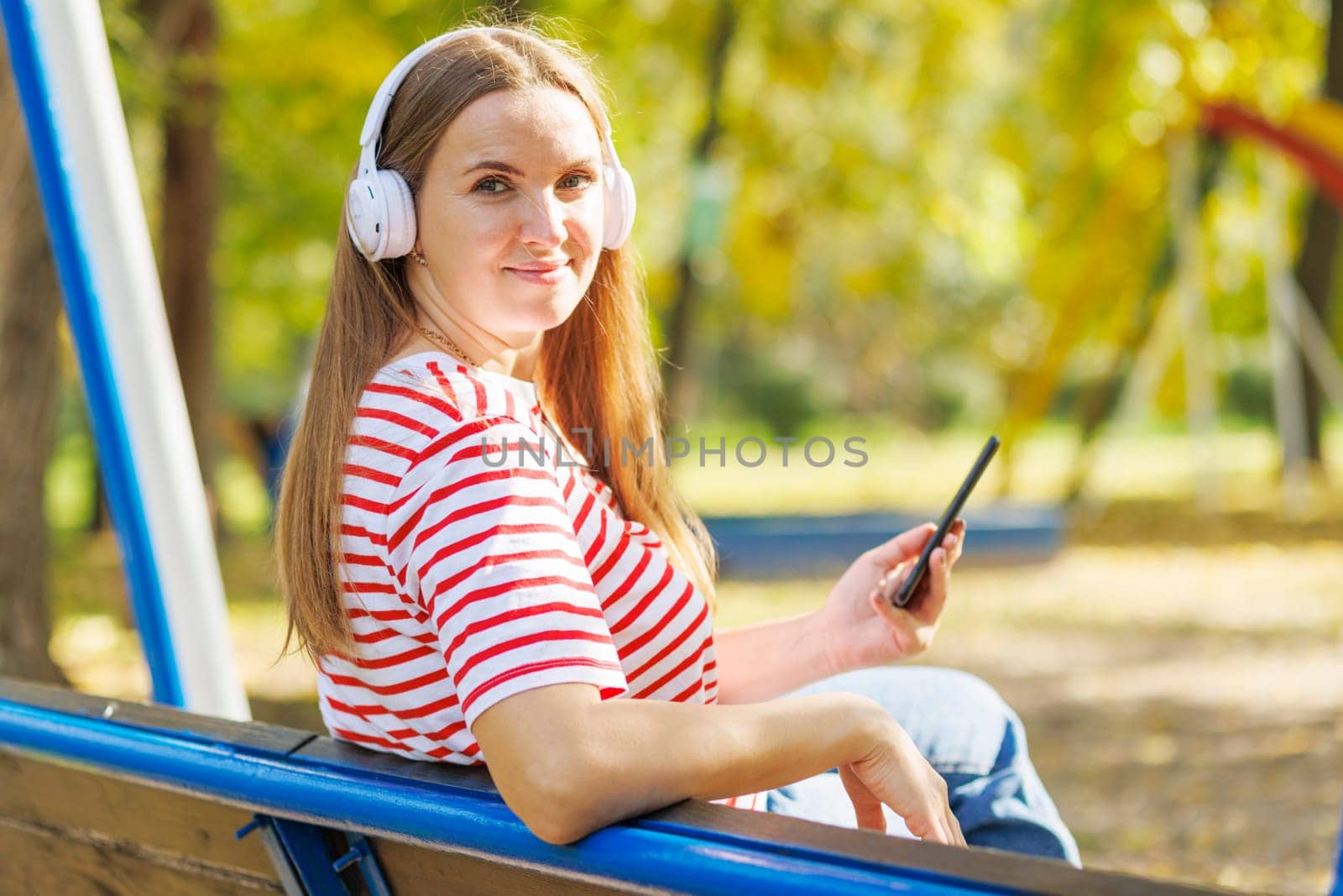 A woman wearing headphones listening to music, breathing fresh air, relaxing while sitting on a bench in sunny day in the park
