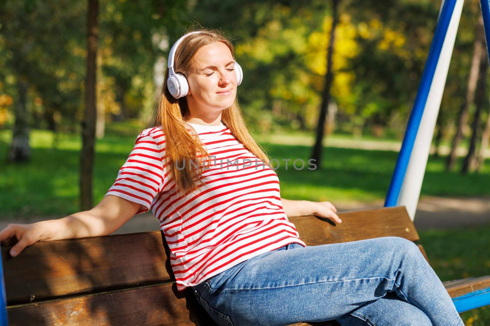 A woman wearing headphones listening to music, breathing fresh air, relaxing while sitting on a bench in sunny day in the park