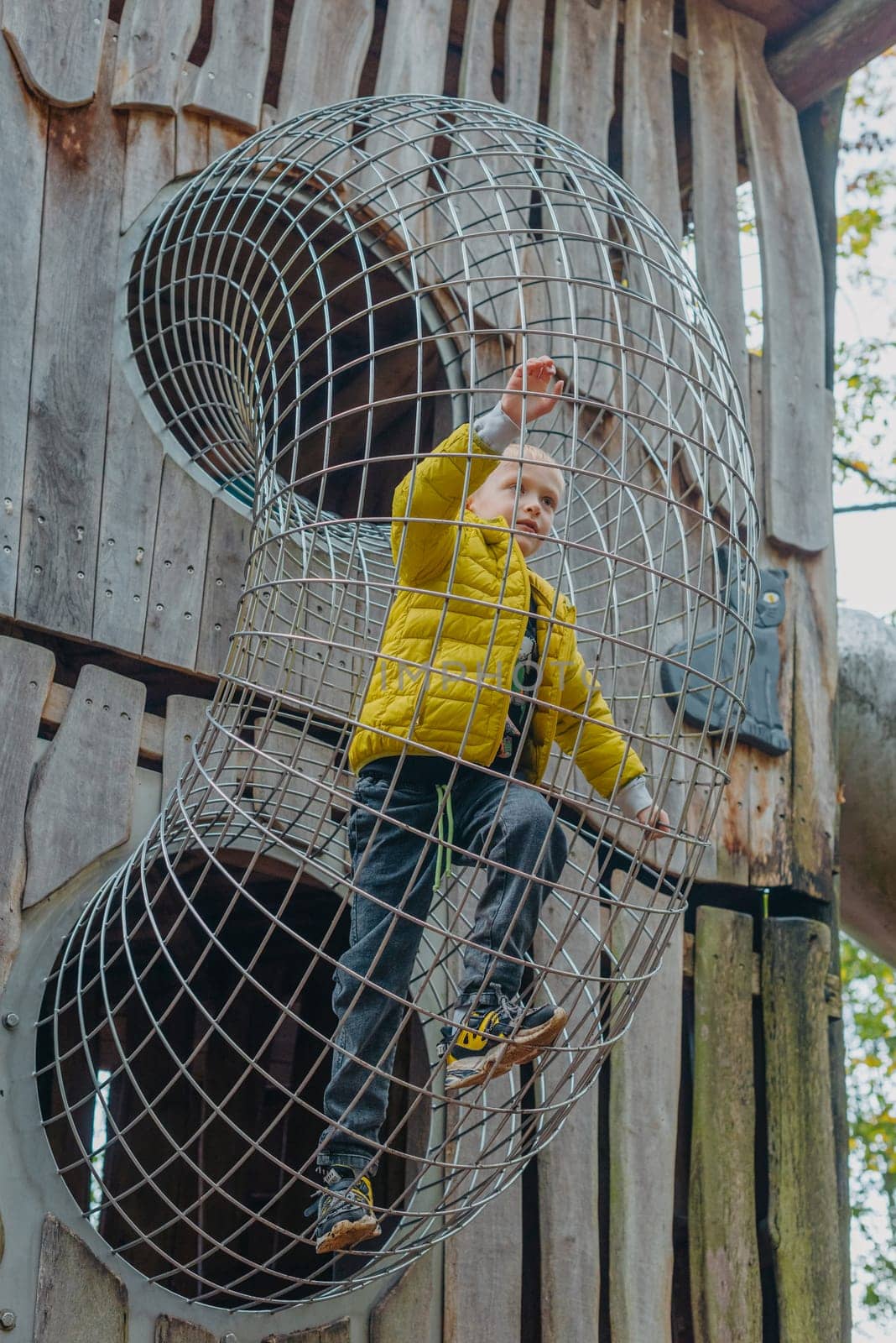 A child climbs up an alpine grid in a park on a playground on a hot summer day. children's playground in a public park, entertainment and recreation for children, mountaineering training. Child playing on outdoor playground. Kids play on school or kindergarten yard. Active kid on colorful slide and swing. Healthy summer activity for children. Little boy climbing outdoors.