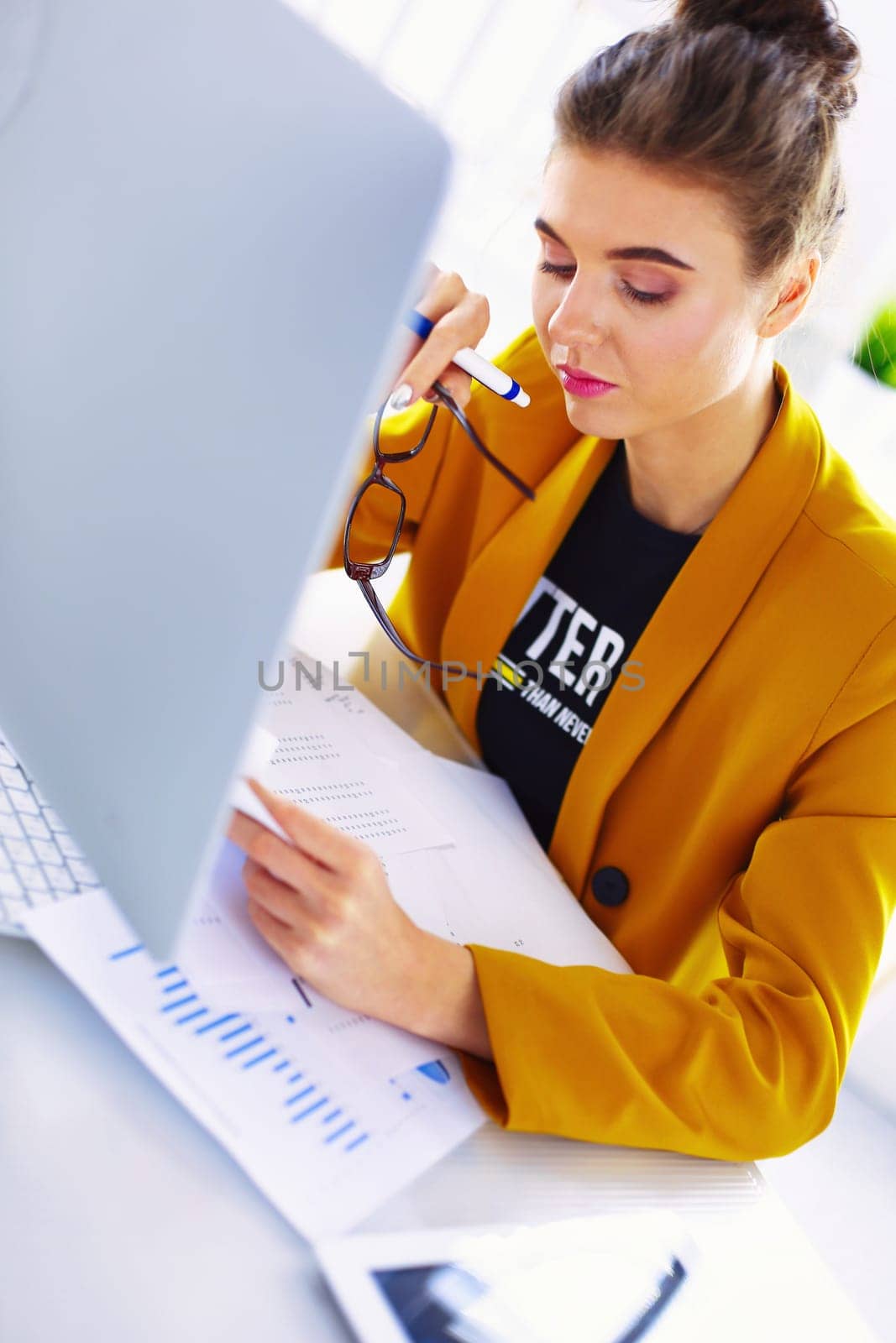 Attractive woman sitting at desk in office, working with laptop computer, holding document.