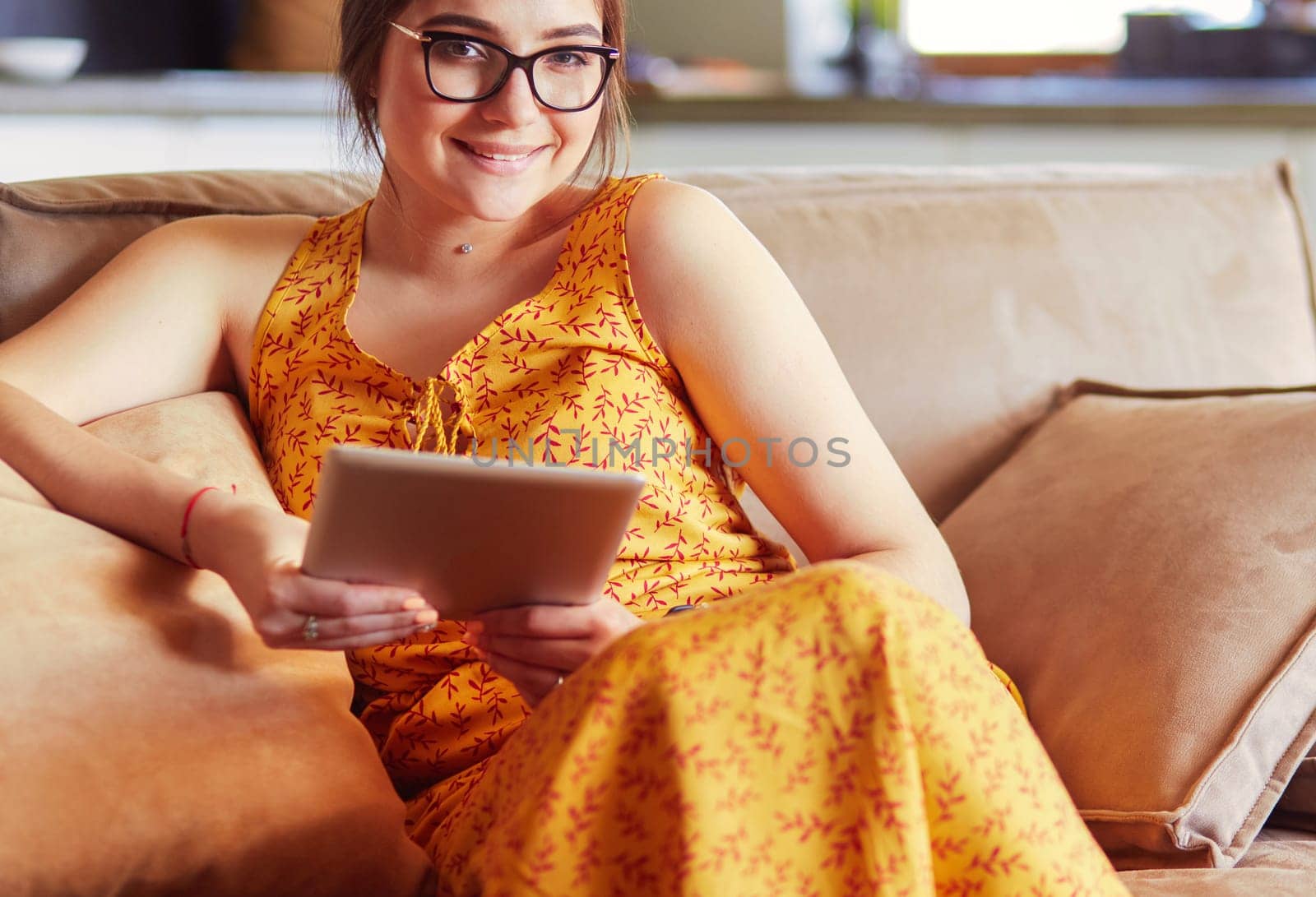 Beautiful young brunette woman at home sitting on sofa or settee using her tablet computer and smiling.