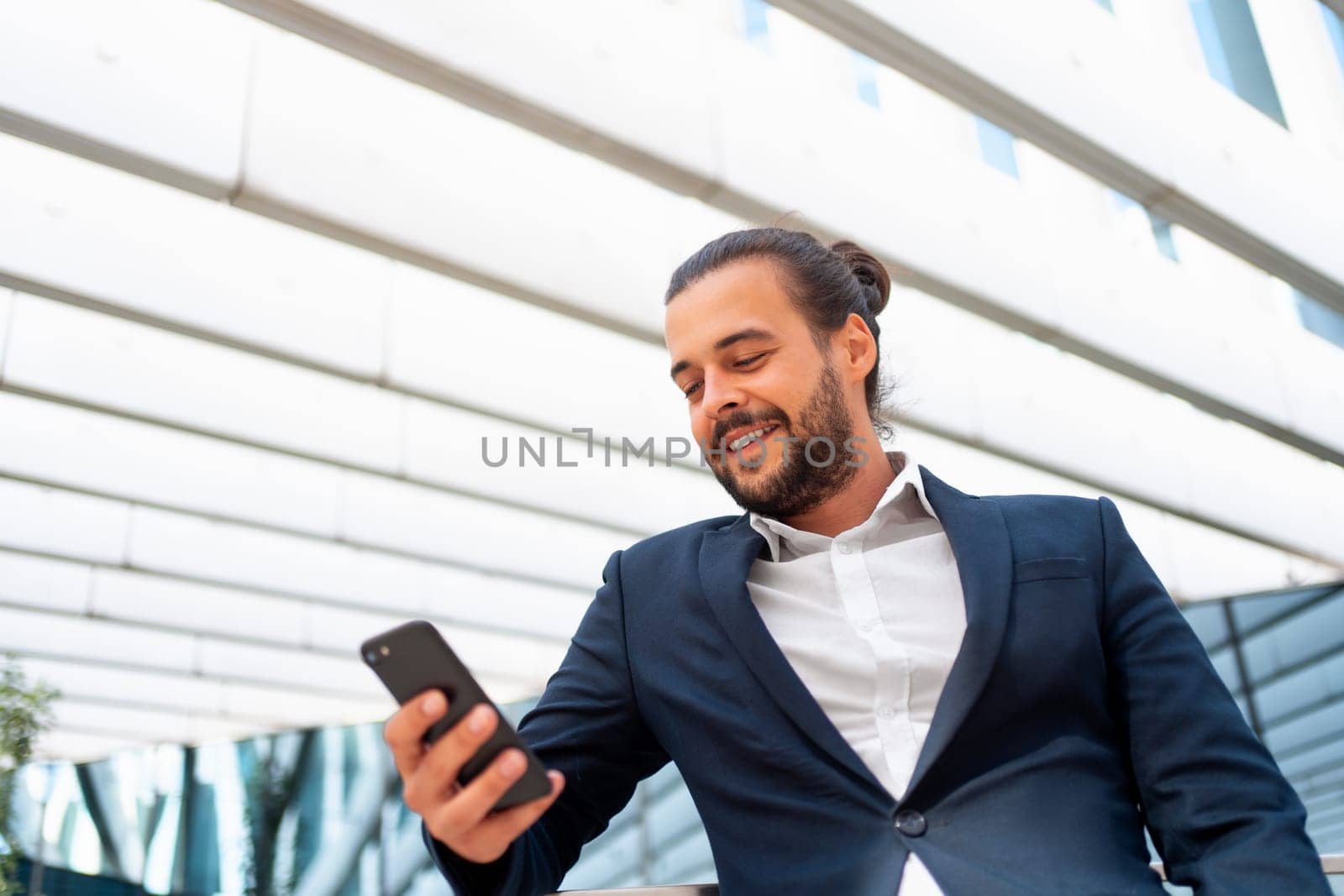 Businessman standing outdoor use smartphone looking device screen and smile. Handsome hispanic male business person in business suit holding smartphone in hand. Modern business person low angle view