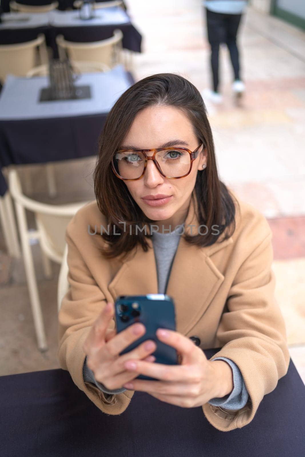 Serious middle-aged woman using smartphone sitting outdoor cafe. Focused businesswoman in glasses dressed trench coat holding smartphone in hand looking at camera. Vertical photo high angle view
