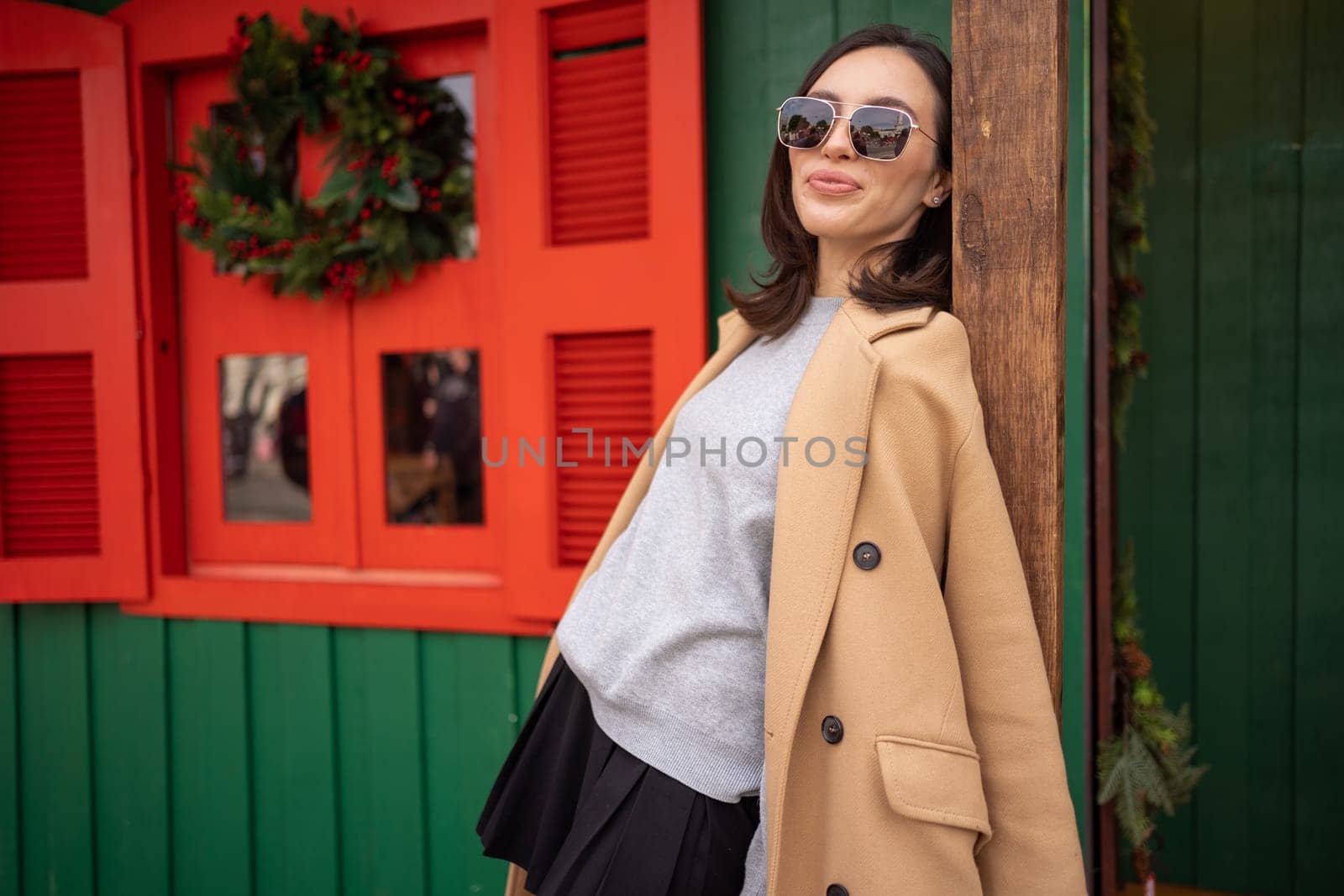 People, season and Christmas concept portrait of happy smiling middle age woman outdoors dressed stylish cream trench coat with Christmas festive decoration on background. Looking at camera
