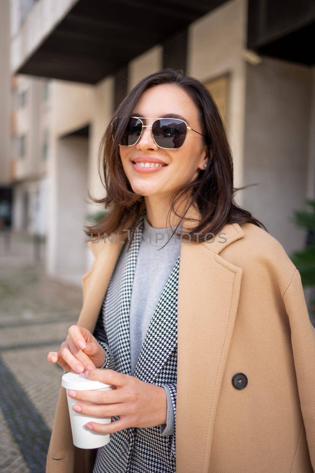 Streetstyle, street fashion concept: woman wearing trendy outfit walking in city. Cream trench coat, sunglasses. Looking camera, vertical photo
