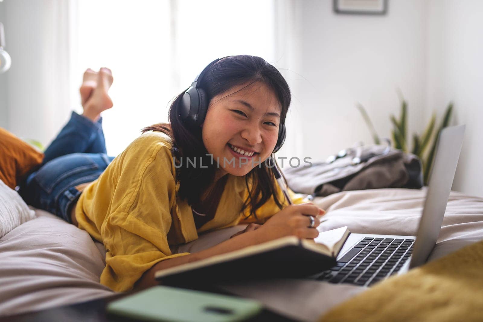 Happy asian teen student doing homework at home bedroom lying on bed looking at camera.Chinese teenage girl using laptop by Hoverstock