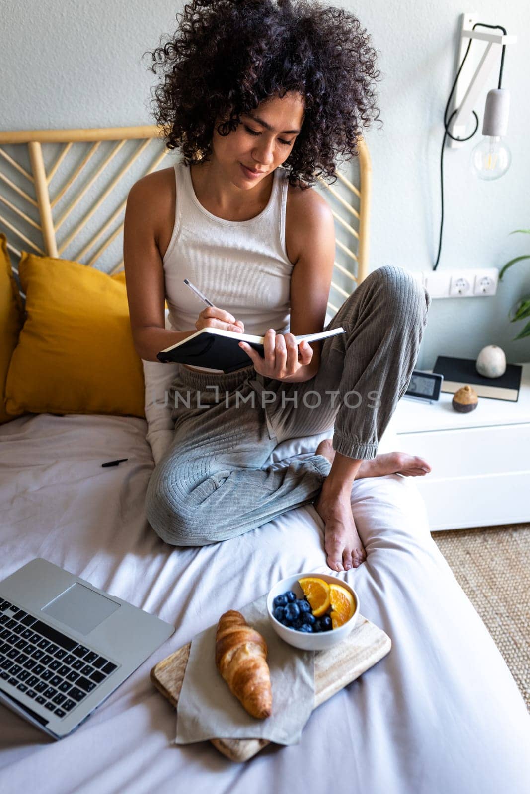 Multiracial latina woman writing daily affirmations on journal while eating breakfast in bed next to laptop.Vertical. by Hoverstock
