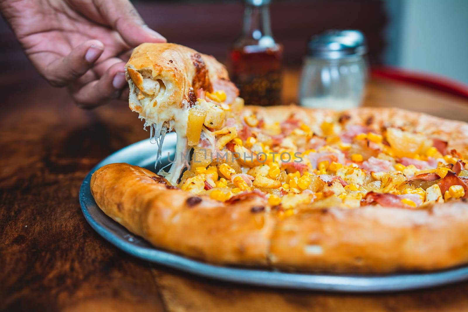 Hand taking a slice of ham and sweetcorn pizza. Homemade ham pizza with cheese and sweet corn served on wooden table by isaiphoto