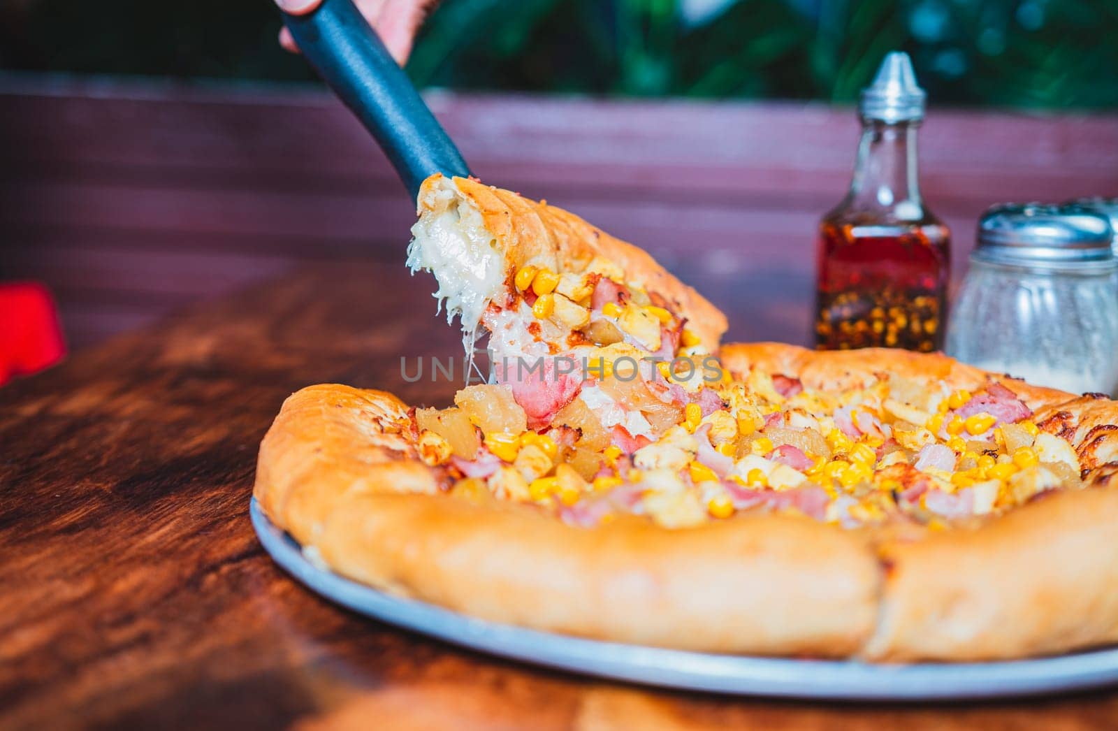 Person hand taking a slice of sweetcorn and ham pizza with spatula. Delicious ham pizza with cheese and sweet corn by isaiphoto