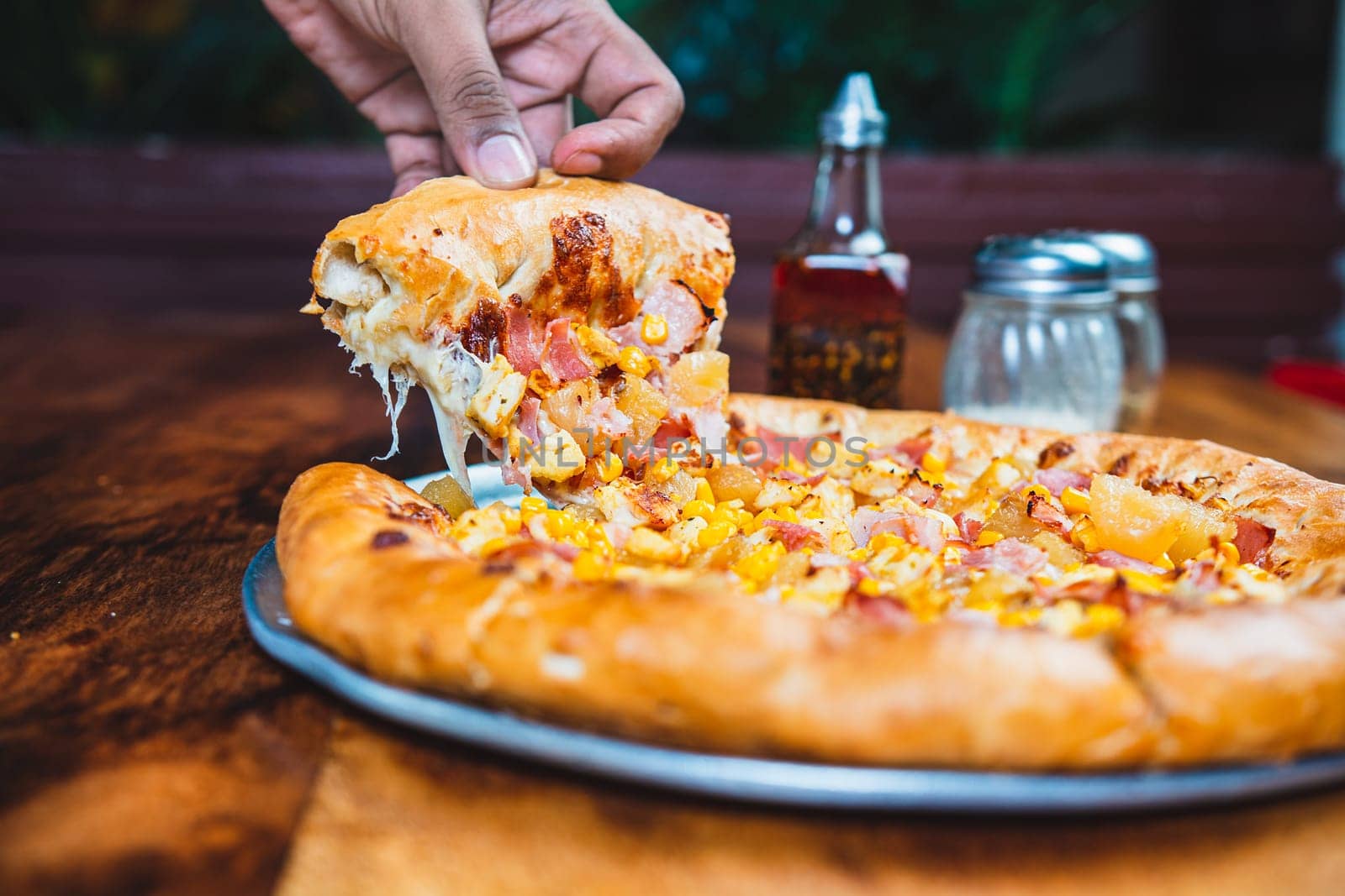 Homemade ham pizza with cheese and sweet corn served on wooden table. Hand taking a slice of ham and sweetcorn pizza