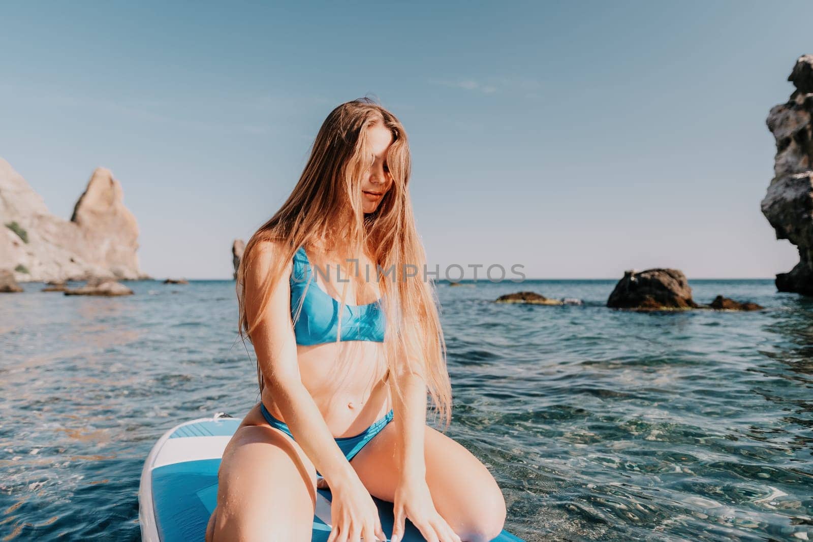 Woman sea sup. Close up portrait of happy young caucasian woman with long hair looking at camera and smiling. Cute woman portrait in a blue bikini posing on sup board in the sea by panophotograph