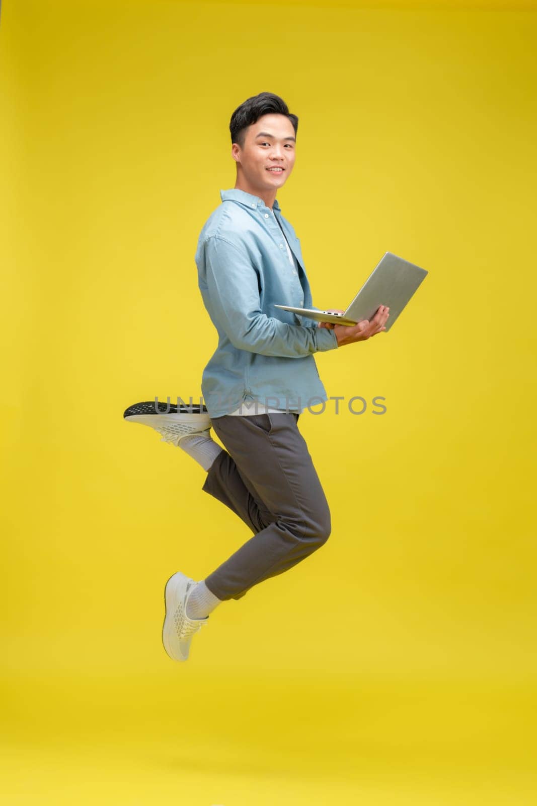 Photo of excited young man jumping isolated over yellow wall background using laptop computer.