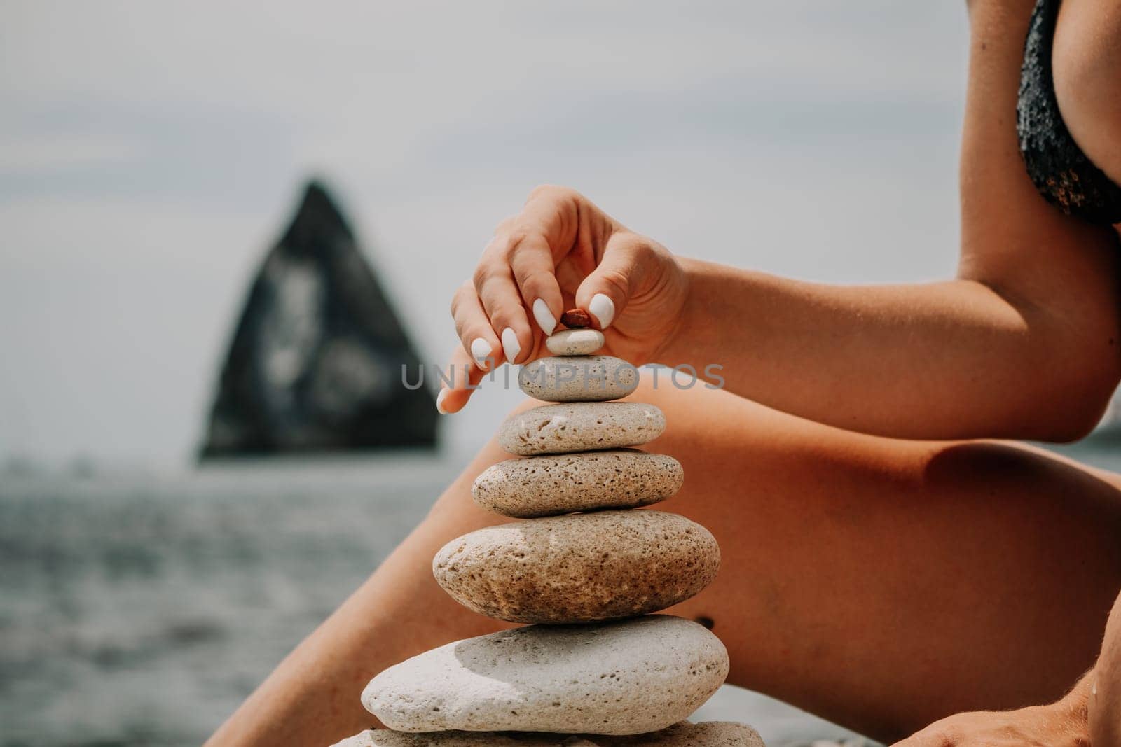 Woman bilds stones pyramid on the seashore on a sunny day on the blue sea background. Happy holidays. Pebble beach, calm sea, travel destination. Concept of happy vacation on the sea, meditation, spa