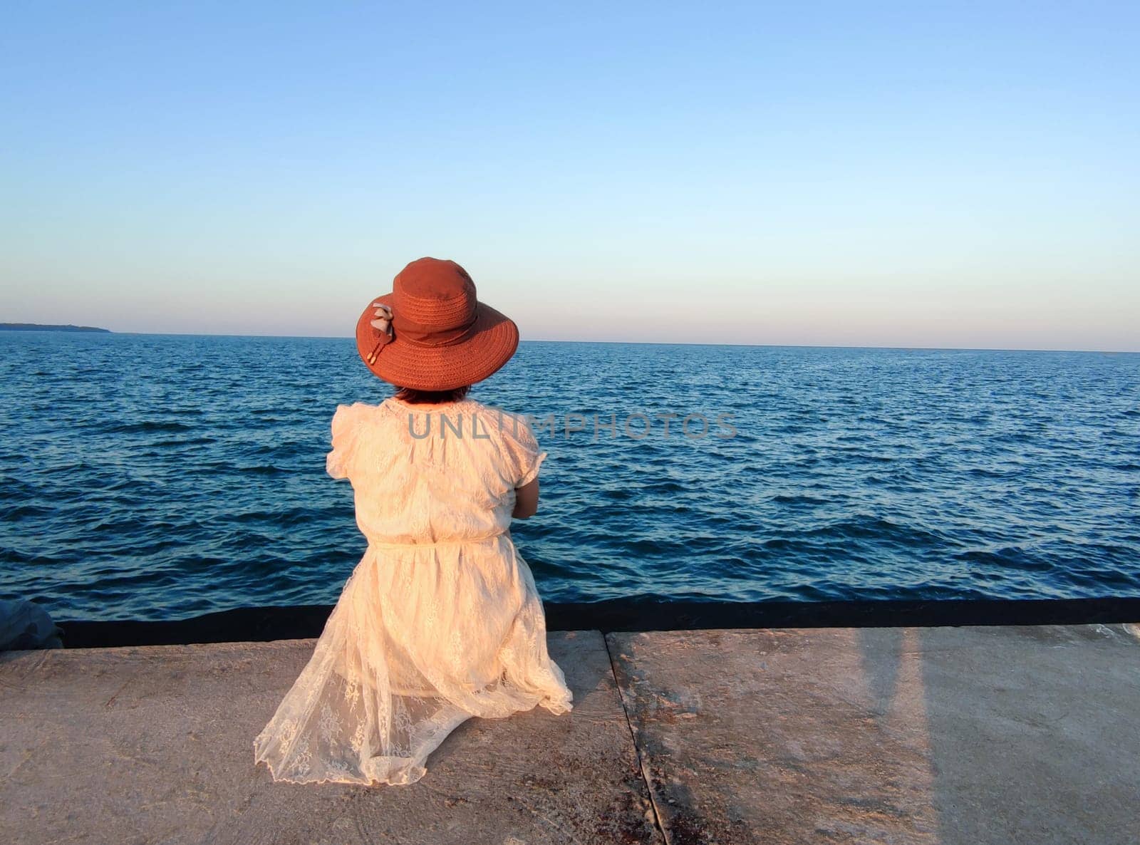 a girl in a white dress and a hat sits on the pier and looks at the sunset sea, rear view by Annado