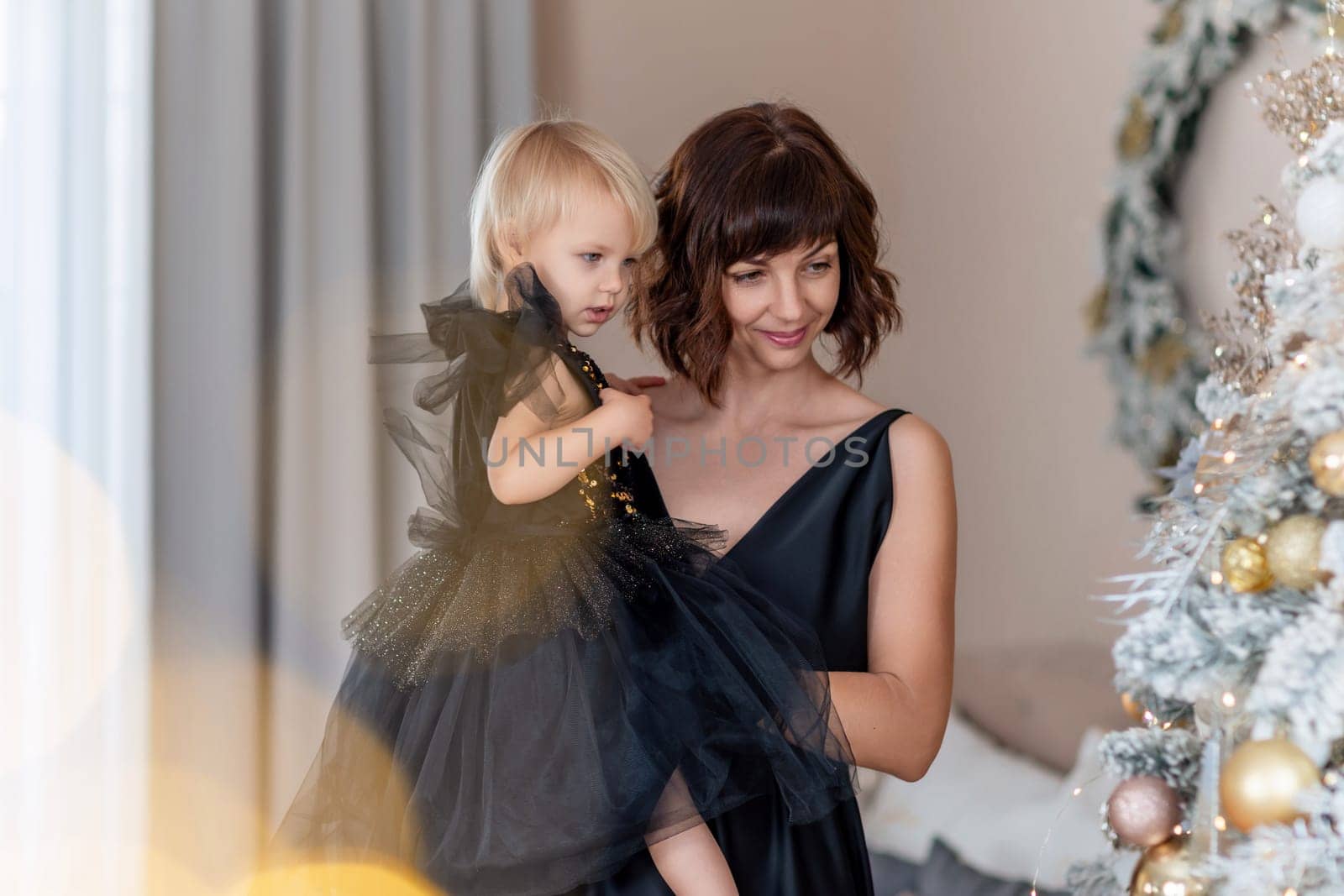 Mother daughter 2 years old Christmas tree. Both are dressed in black dresses, the mother holds the girl in her arms and both look at each other. The family celebrates Christmas