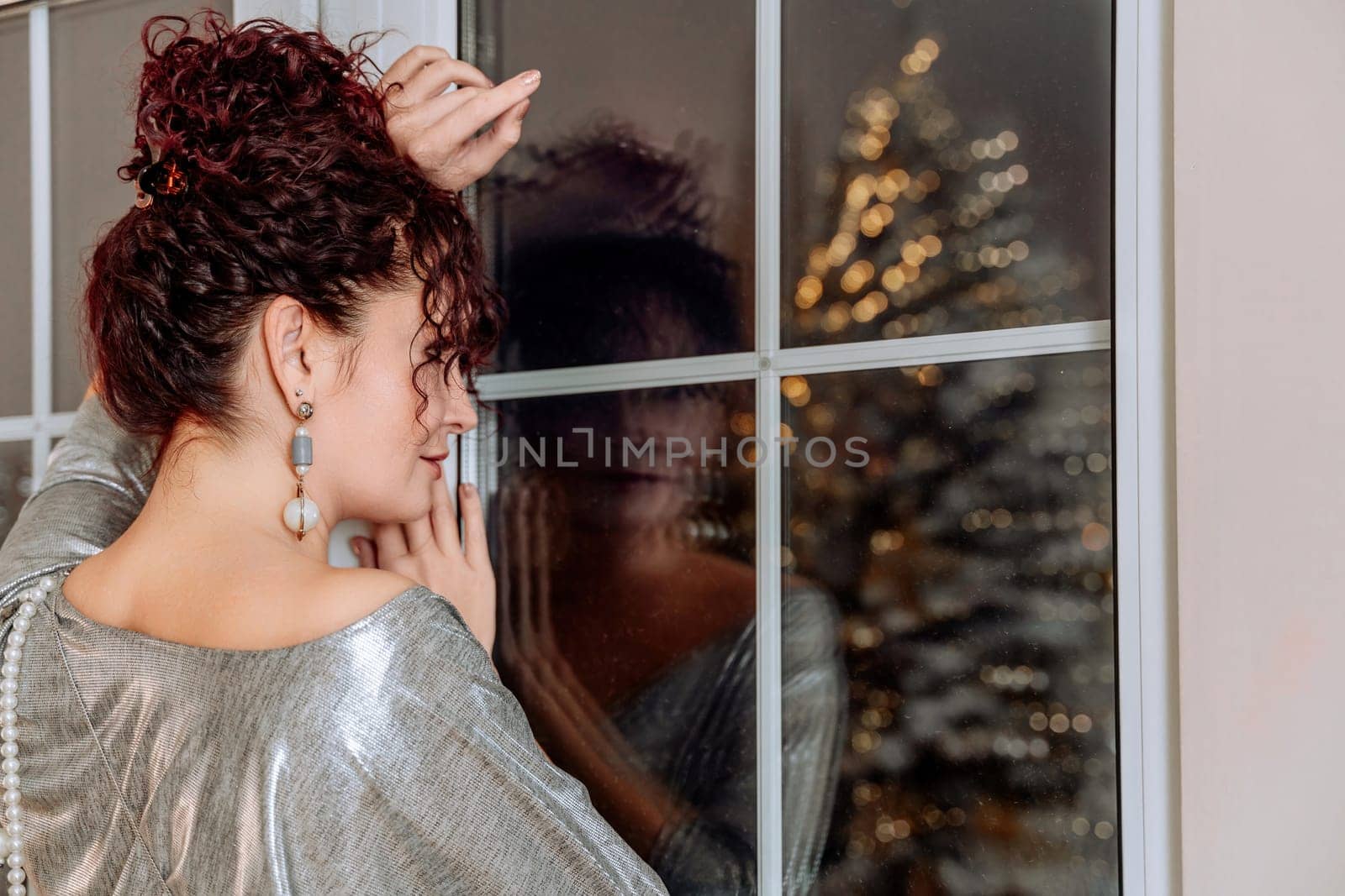 Woman portrait window bokeh christmas tree. A curly-haired middle-aged woman looks out of a dark window where the bokeh of garlands from the Christmas tree is reflected. She is dressed in a silver dress with pearl beads and earrings