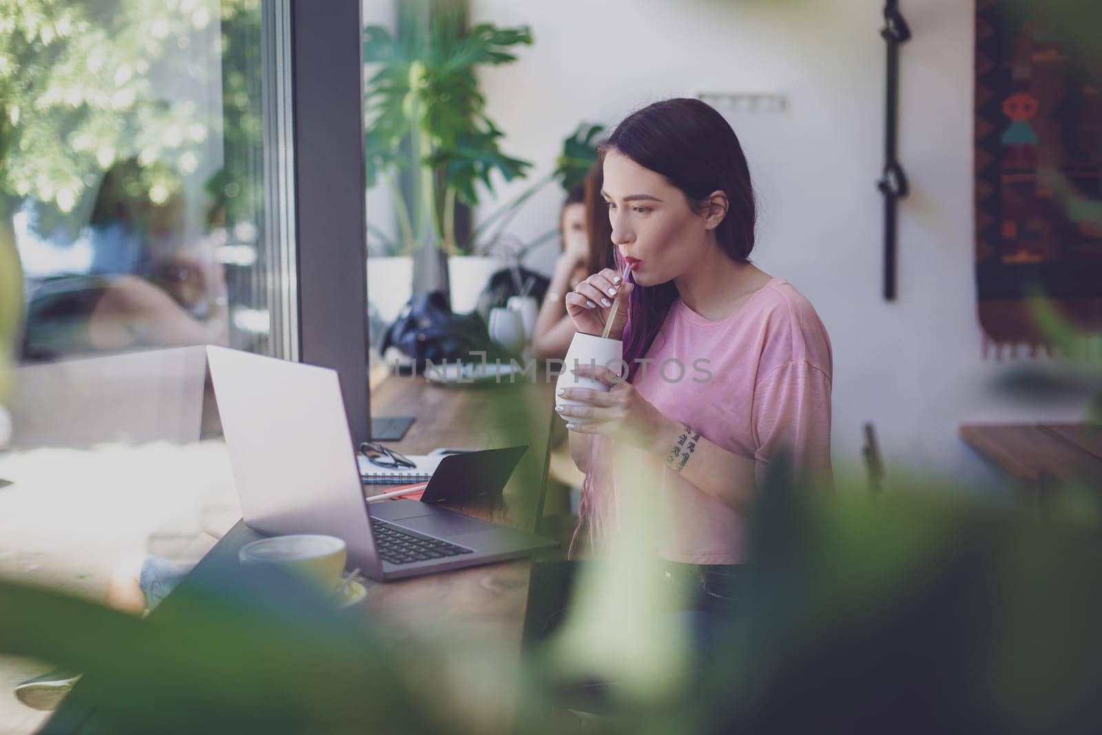 Side view of young businesswoman sitting at table in coffee shop. On table cup of coffee and laptop. In background white wall and window. Girl shopping online.