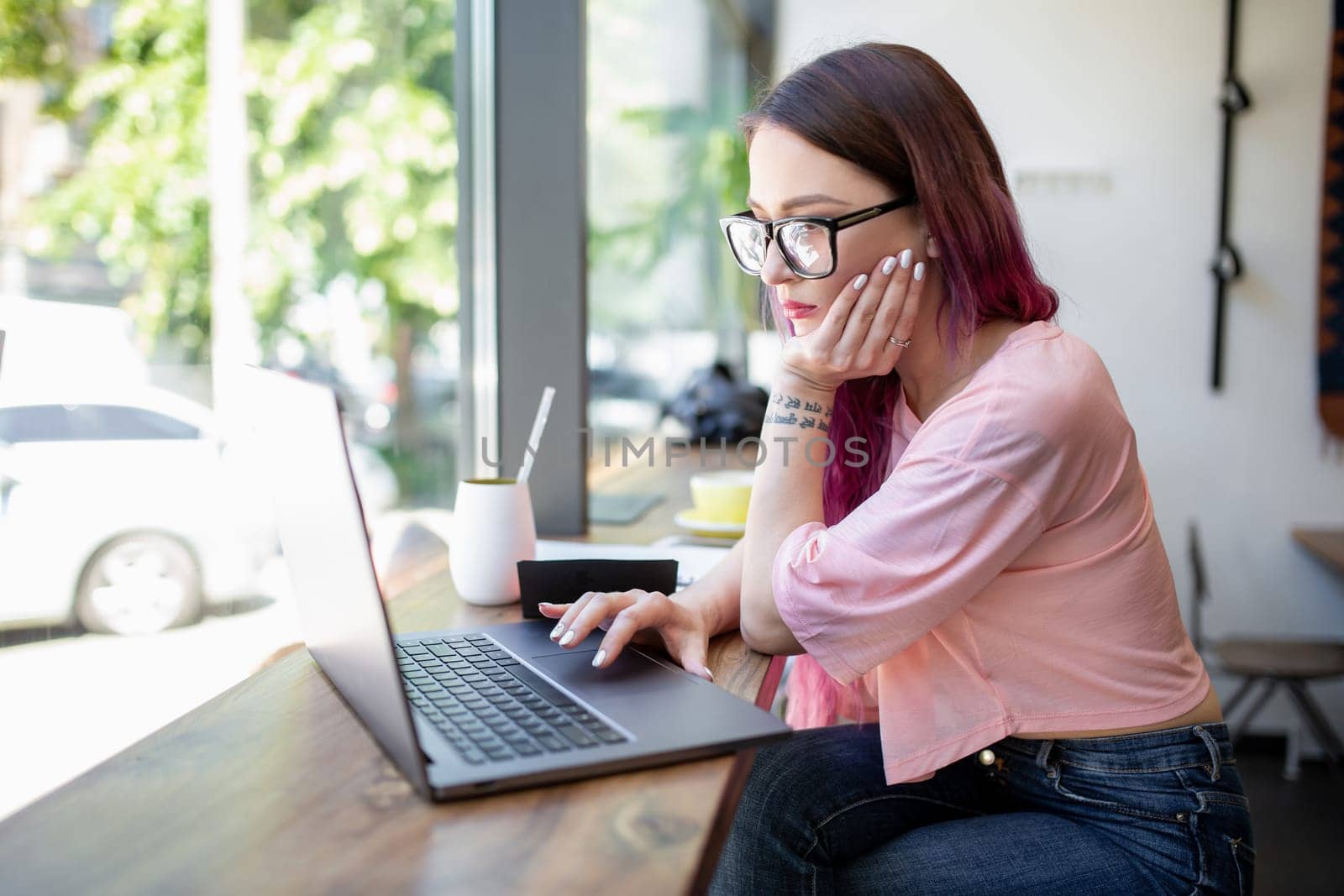 Side view of young businesswoman sitting at table in coffee shop. On table cup of coffee and laptop. In background white wall and window. Girl shopping online.
