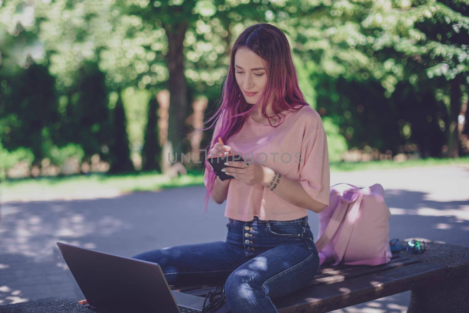 Woman writing in a notebook sitting on a wooden bench in the park. Girl working outdoors on portable computer, copy space. Technology, communication, freelance and remote working concept.
