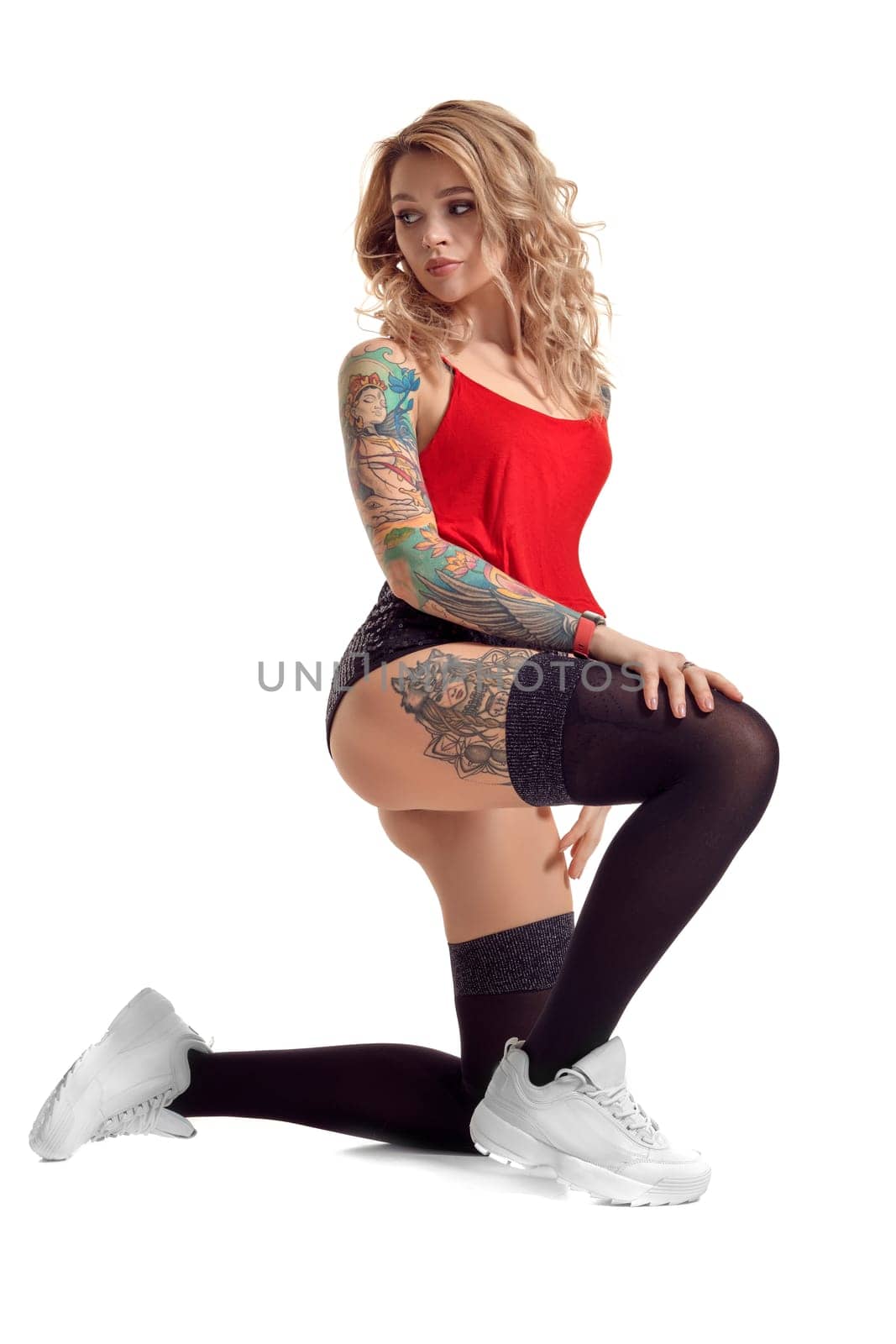 Sexy blond female with tattoed body and long curly hair is looking away while posing isolated on white background with copy space. Young woman wearing in a black stockings and mini shorts, red top and white sneakers. Booty twerk dance in studio.