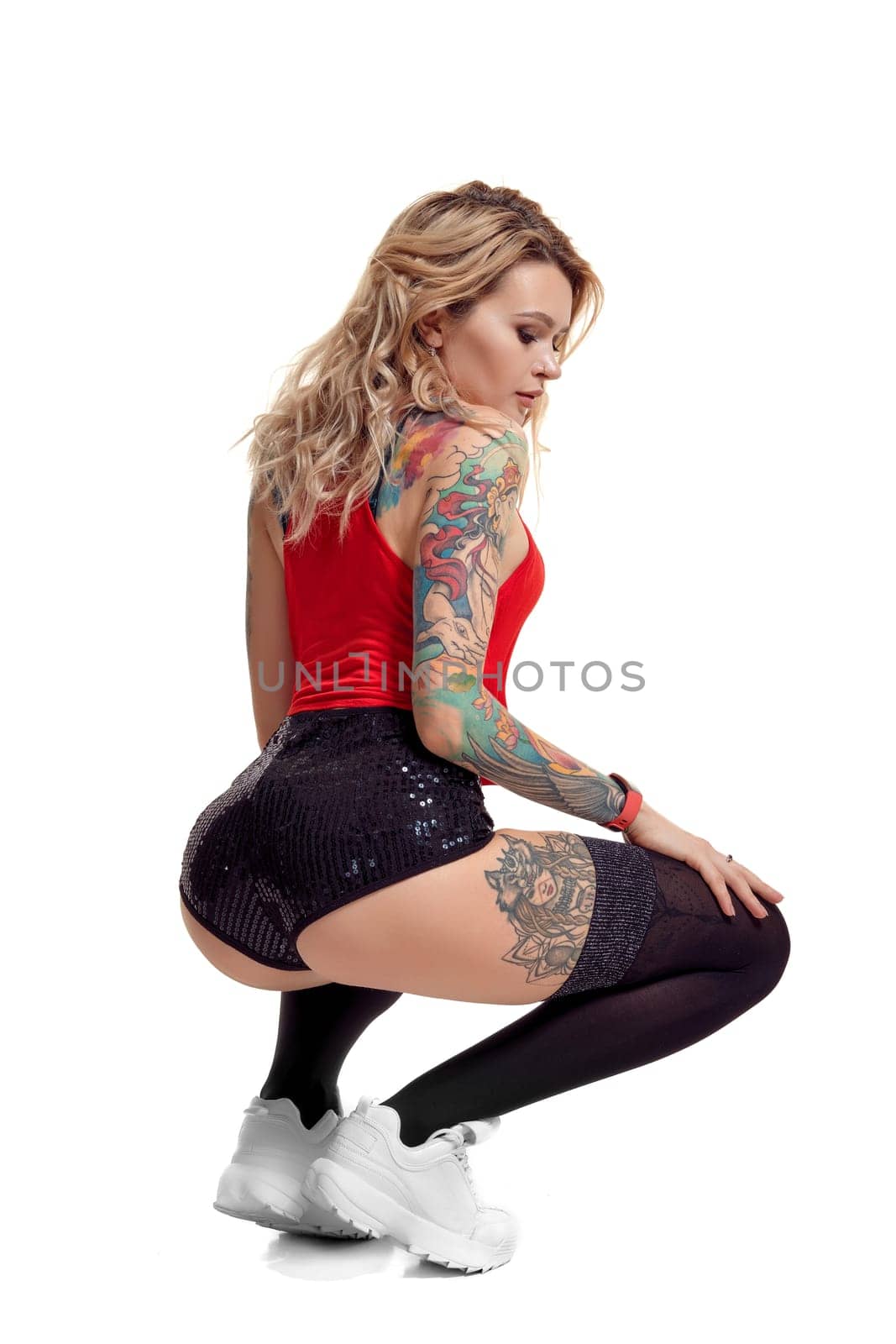 Sexy blond lady with tattoed body and long curly hair is posing back, isolated on white background with copy space. Young woman wearing in a black stockings and mini shorts, red top and white sneakers. Booty twerk dance in studio.