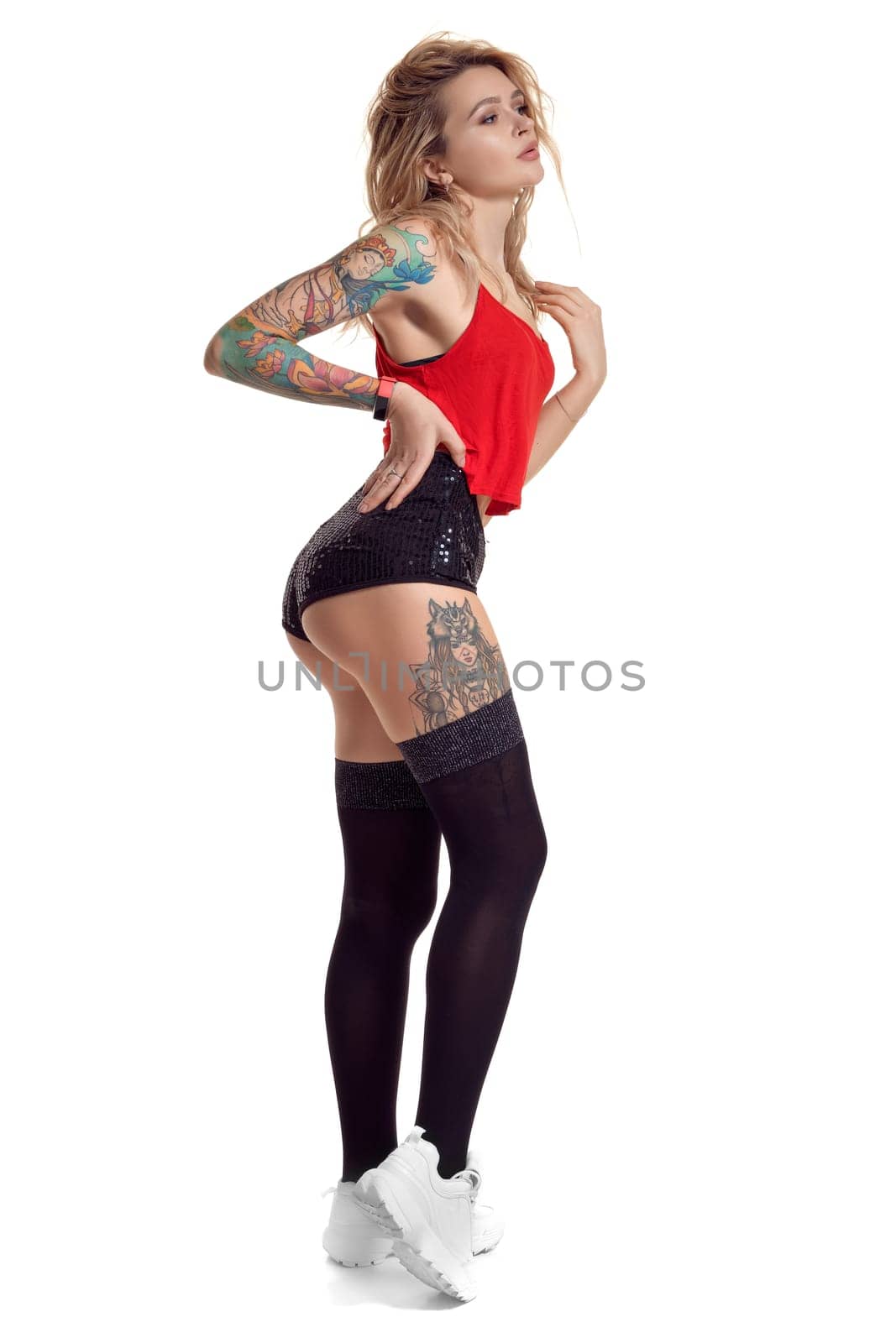 Sexy blond girl with tattoed body and long curly hair is posing sideways isolated on white background with copy space. Young woman wearing in a black stockings and mini shorts, red top and white sneakers. Booty twerk dance in studio.