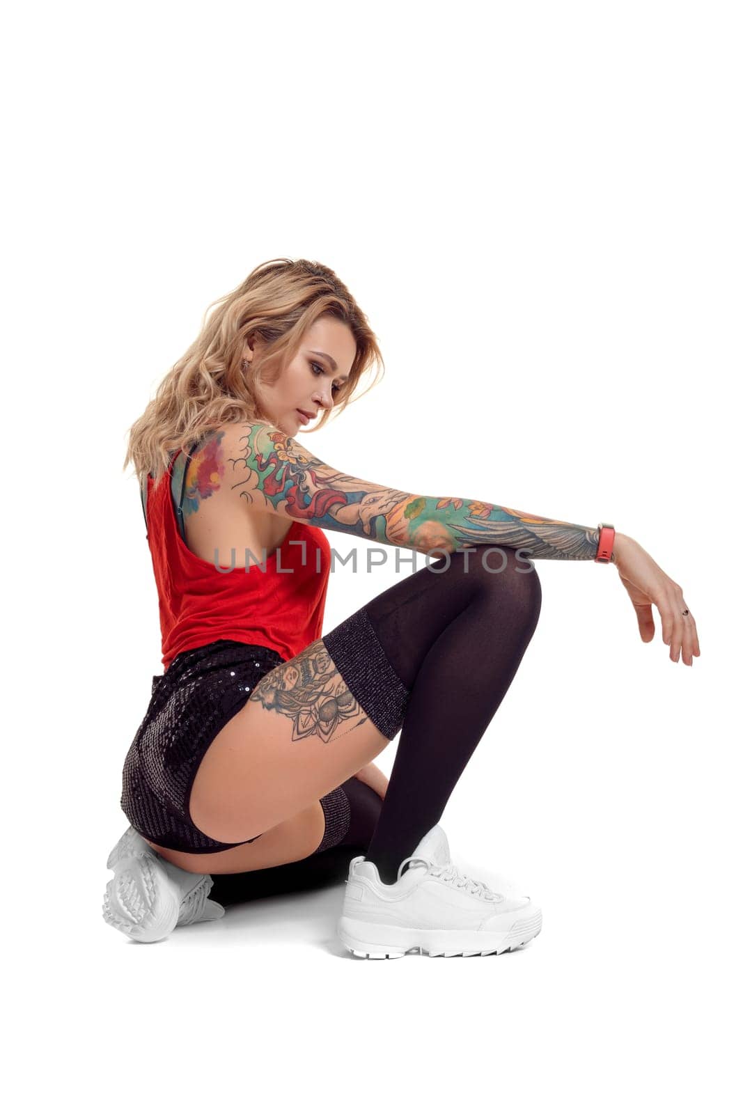 Charming blond woman with tattoed body and long curly hair is posing sitting sideways, isolated on white background with copy space. Young girl wearing in a black stockings and mini shorts, red top and white sneakers. Booty twerk dance in studio.