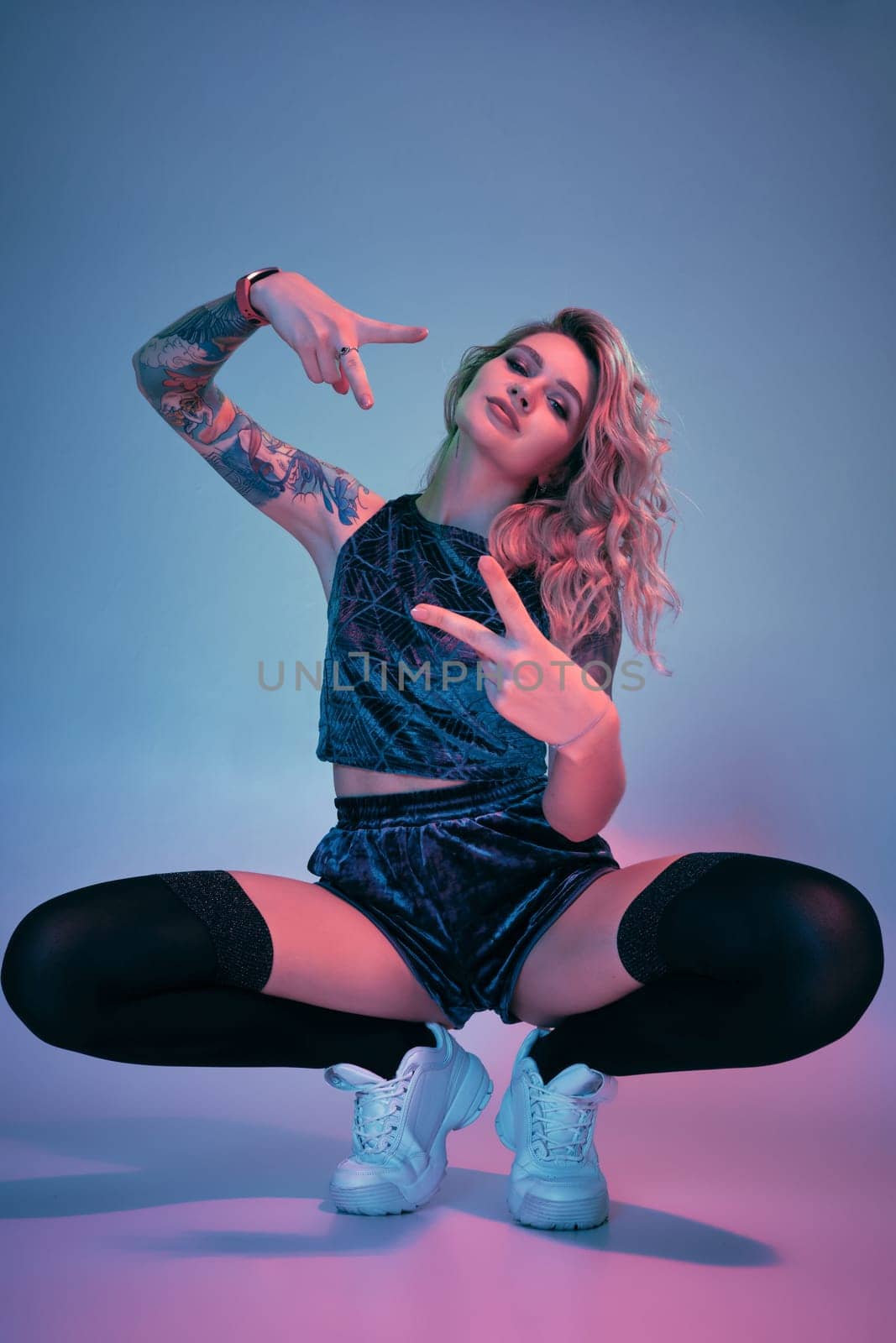 Beautiful young blonde tattoed girl in blue velour booty shorts and t-shirt, black stockings, squats in front of the camera in a confident pose. Pink and blue background