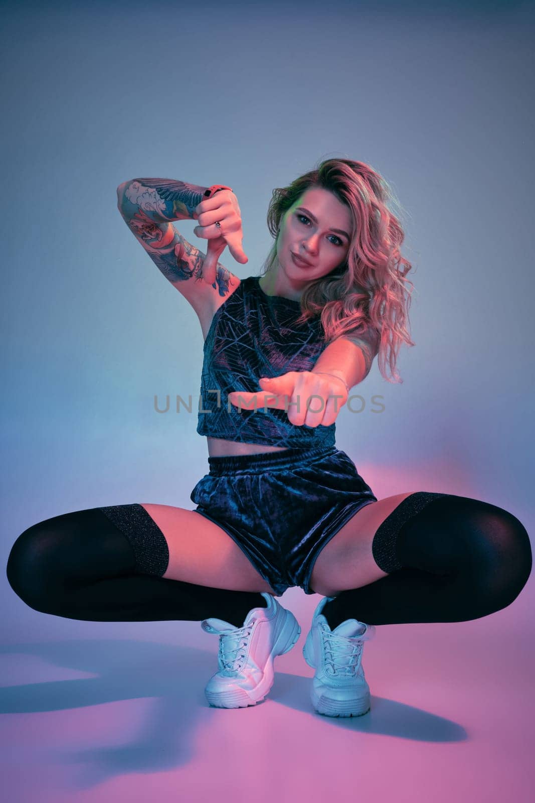 Beautiful young blonde tattoed girl in blue velour booty shorts and t-shirt, black stockings, squats in front of the camera in a confident pose. Pink and blue background