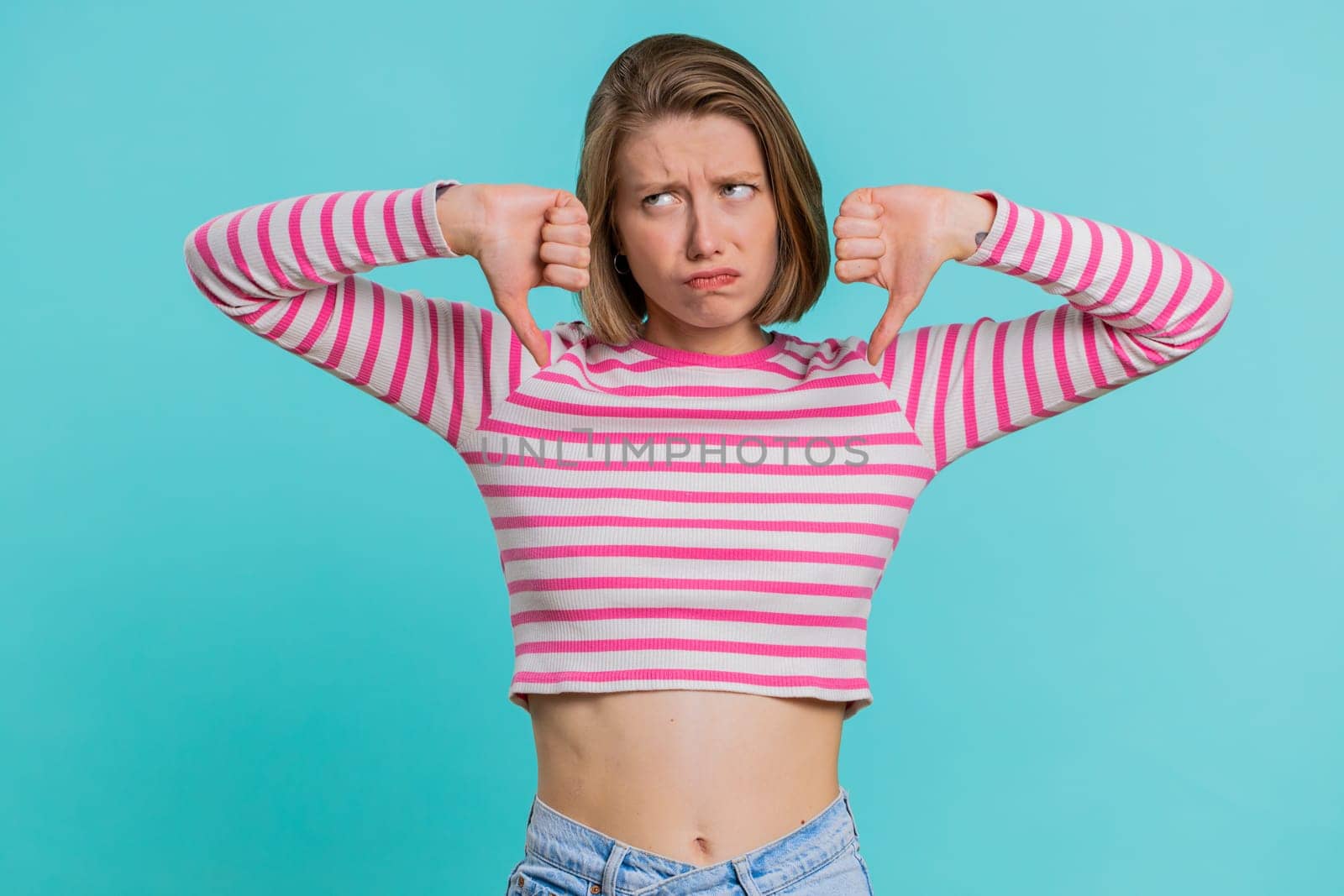 Dislike. Upset unhappy young woman showing thumbs down sign gesture, expressing discontent, disapproval, dissatisfied, dislike. Pretty girl in crop top. Indoors isolated on blue studio background