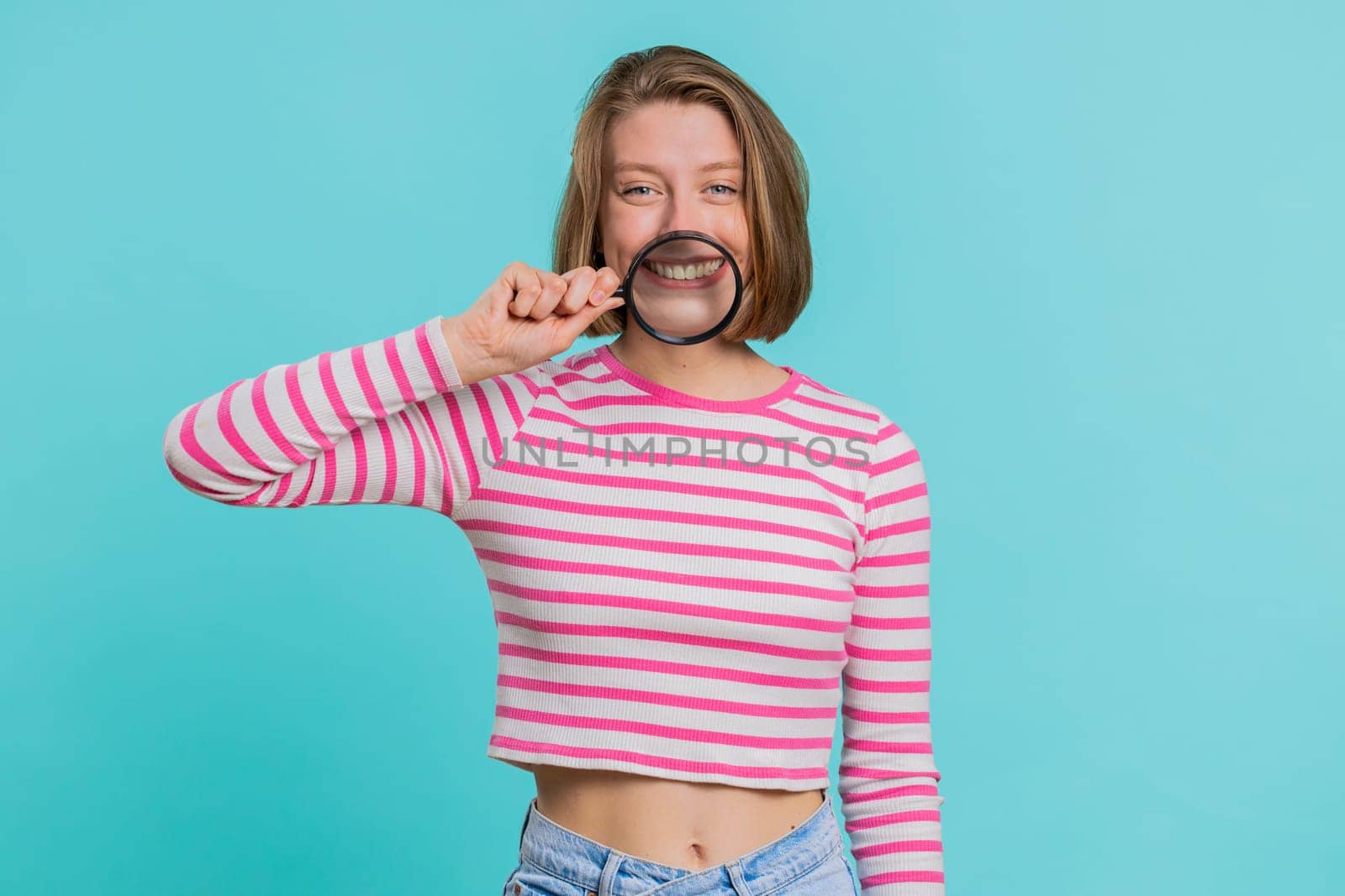 Young woman holding magnifier glass loupe on healthy white teeth, looking at camera with happy expression, showing funny silly face smiling mouth. Girl in crop top isolated on blue studio background