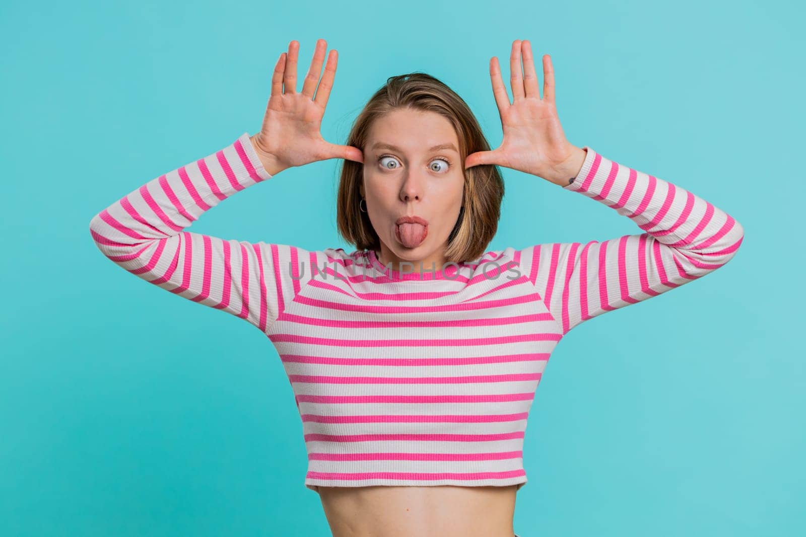 Funny comical playful young woman making silly facial expressions and grimacing, fooling around, showing tongue, idiotic expression. Pretty girl in crop top isolated alone on blue studio background