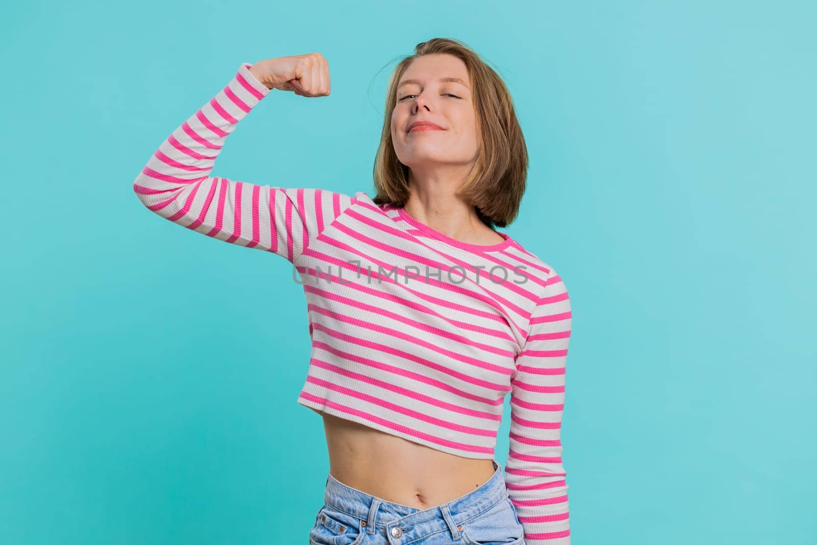 I am strong and independent. Young woman showing biceps and looking confident, feeling power strength to fight for rights, energy to gain success win. Girl isolated alone on blue studio background