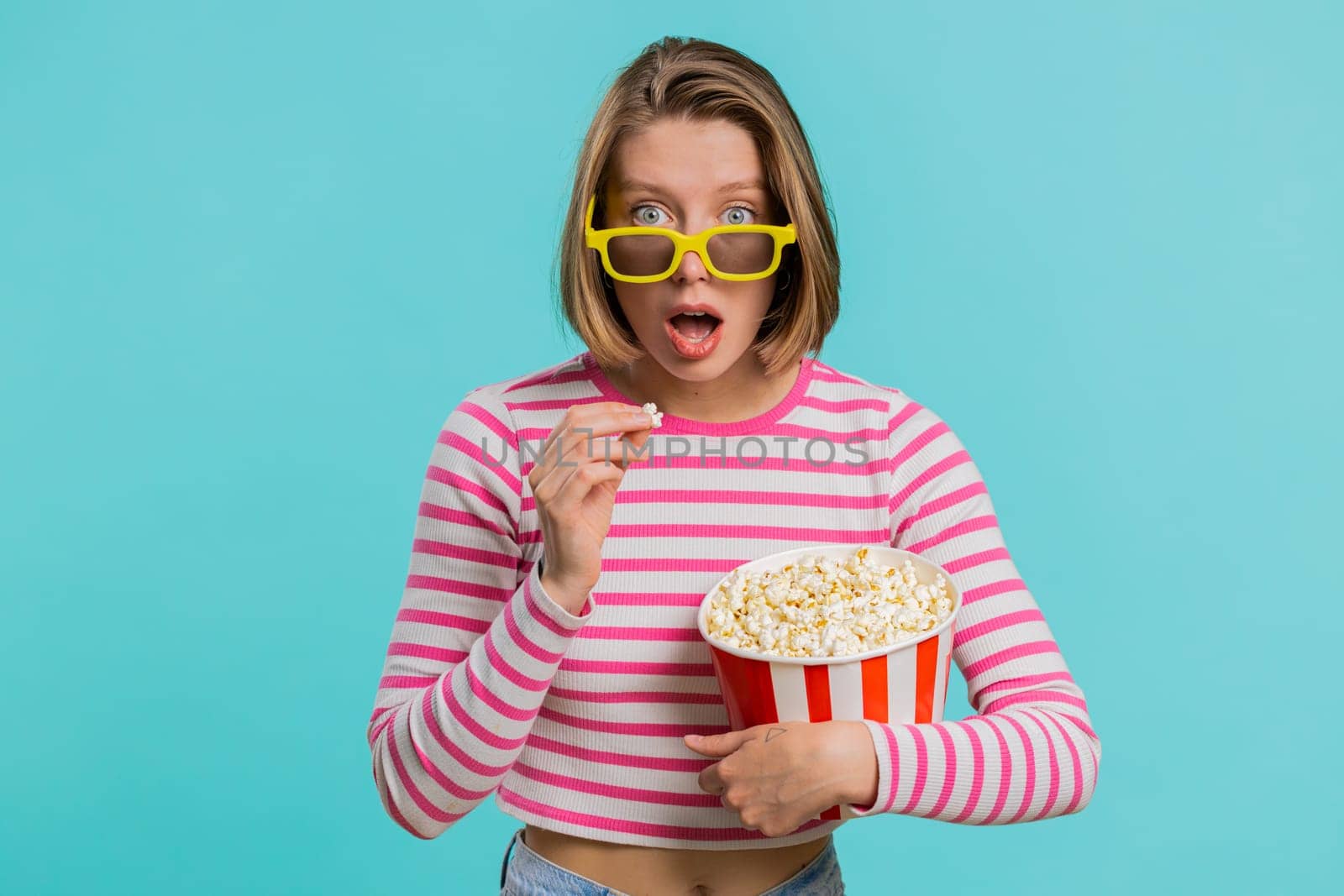 Excited young woman in 3D glasses eating popcorn and watching interesting tv serial, sport game, film, online social media movie content. Lovely girl in crop top isolated on blue studio background