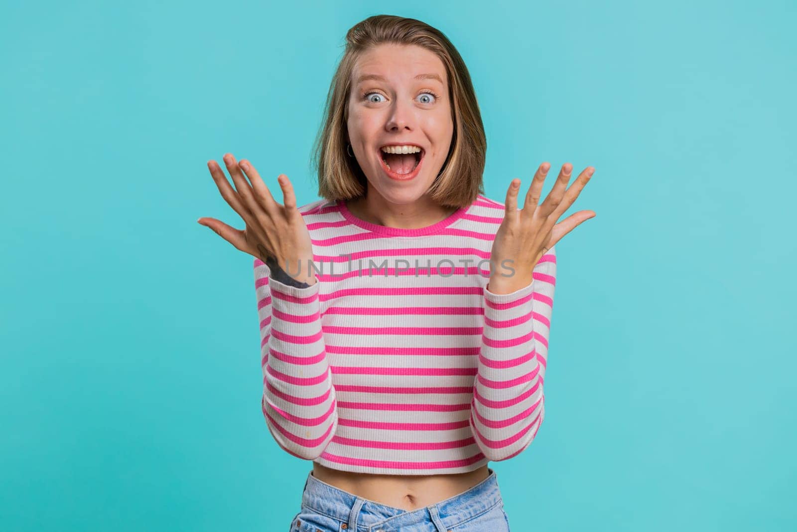 Oh my God, Wow. Excited amazed winner woman raising hands in surprise looking at camera with big eyes, shocked by sudden victory. Sincere girl in crop top isolated alone on blue studio background