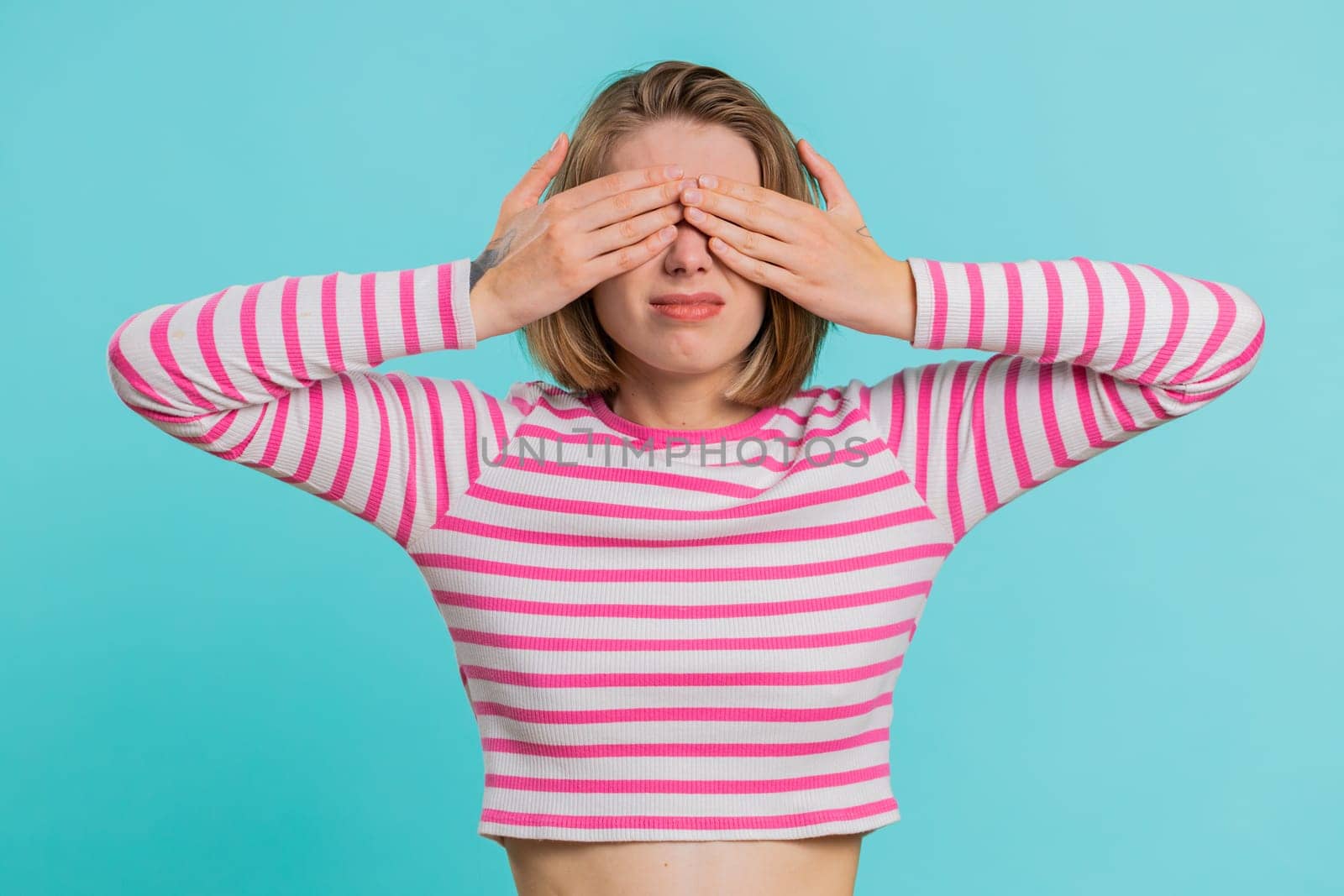 Dont want to look at this, awful. Afraid woman in crop top closing eyes with hand showing stop gesture, confused shy scared to watch. Young girl isolated on blue studio background. People lifestyles