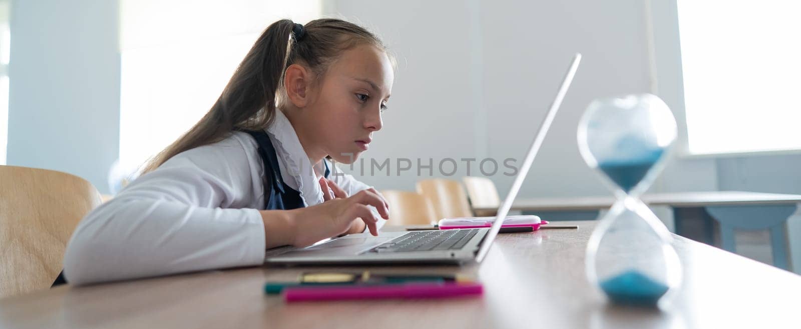Caucasian girl sits at a desk at school and studies at a laptop. Hourglass on the table. by mrwed54