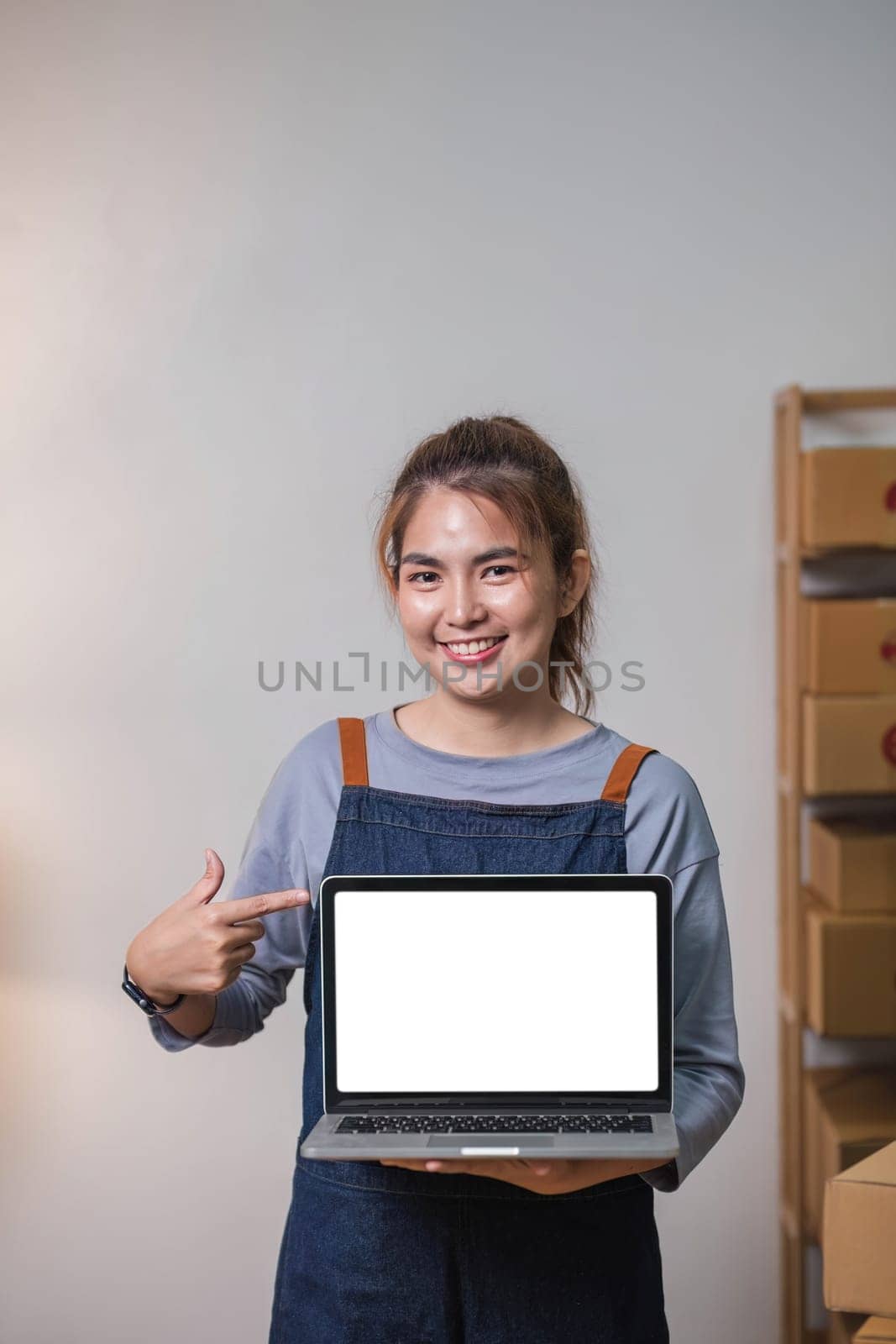 Asian SME business woman showing laptop empty screen at home office. Portrait young Asian small business owner home office, online sell marketing delivery by wichayada