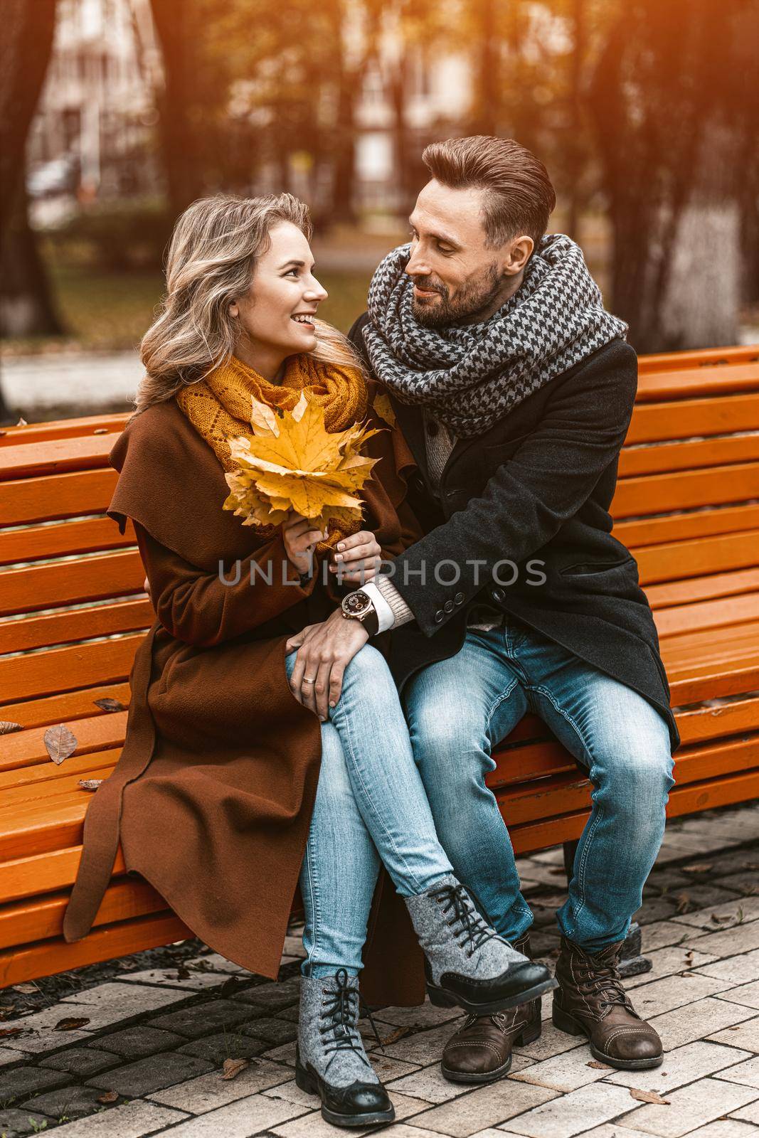 Front view of a couple in love sitting on the bench hugged in park wearing coats and scarfs collecting a bouquet of fallen leaves. Love story concept. Tinted image. 