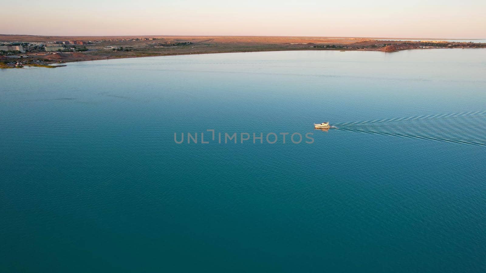 The boat sails in the bay at sunset near the city. In the distance, you can see the pipes of the plant and the orange sun. Ecology of Lake Balkhash. Catamarans float, city lights are lit. Kazakhstan