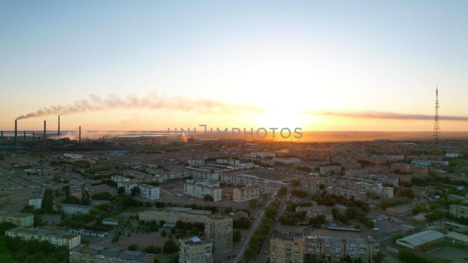 An epic sunset with a view of the smoking factory and the city. The embankment of Lake Balkhash. Low houses are standing and there are few trees. The pipes from the factory are smoking. Kazakhstan