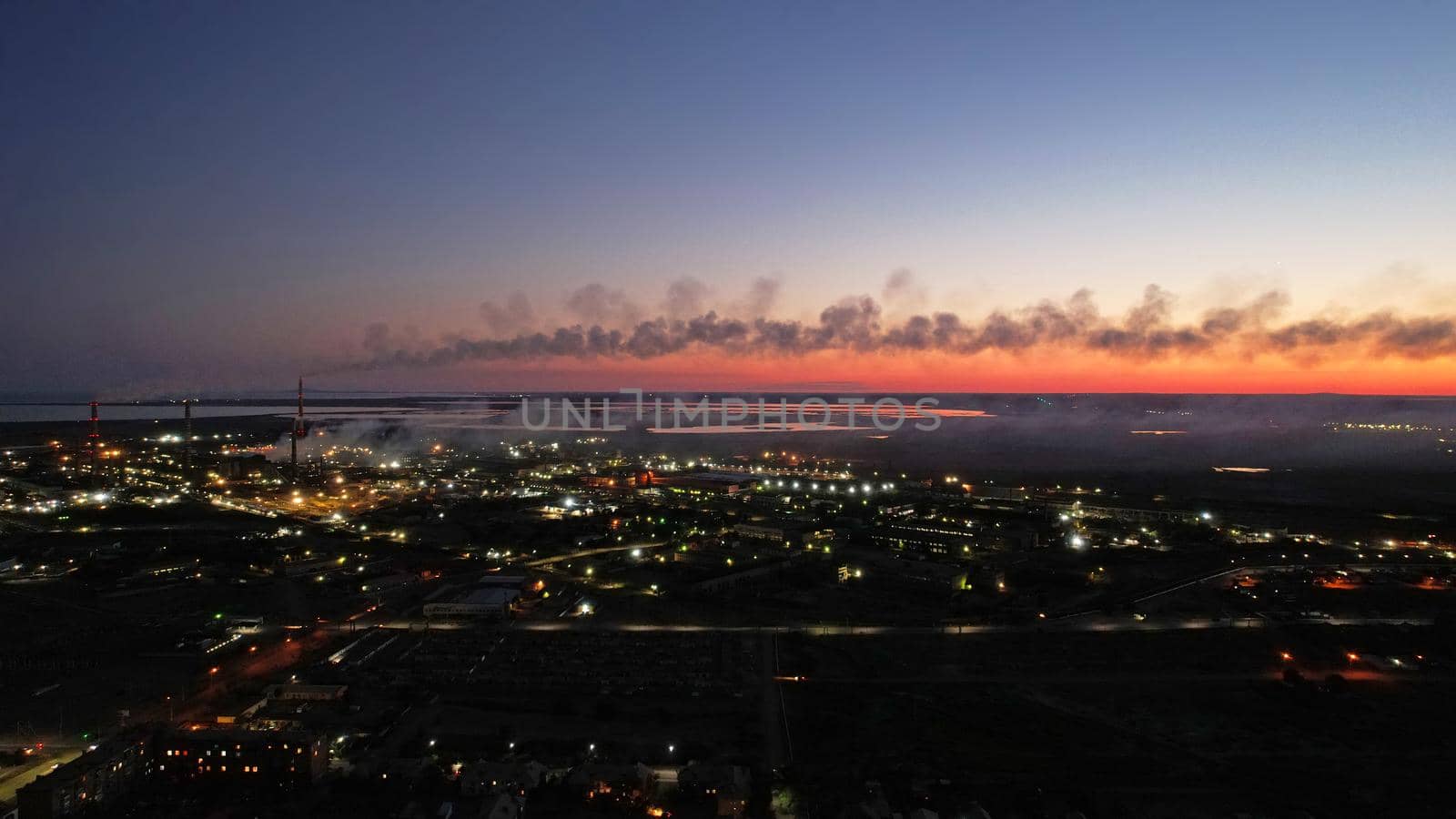 An epic sunset with a view of the smoking factory and the city. The embankment of Lake Balkhash. Low houses are standing and there are few trees. The pipes from the factory are smoking. Kazakhstan