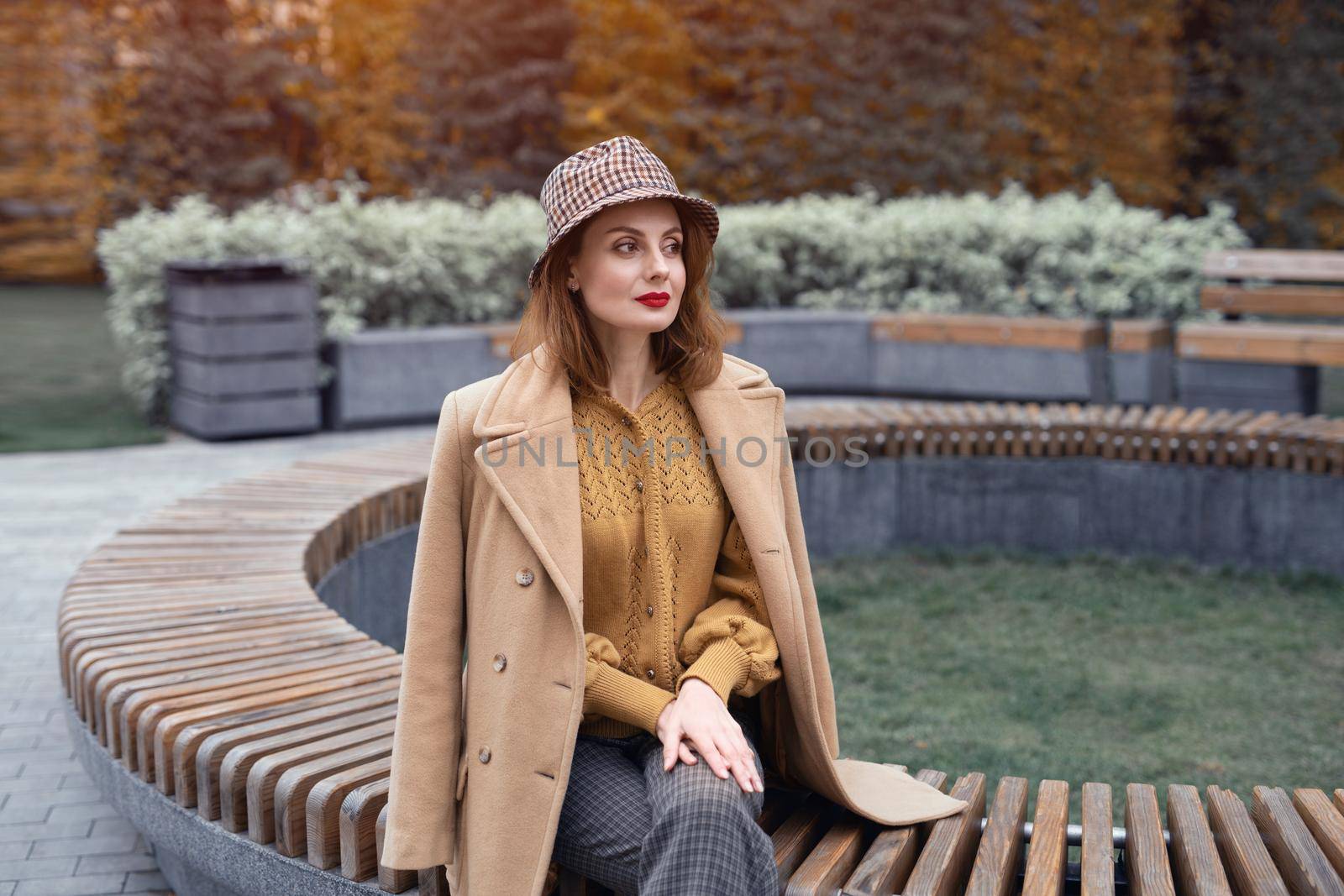 Charming young woman in beige coat and plaid hat sits on round bench waiting for her date or girlfriends. Autumn walk concept. Tinted photo.