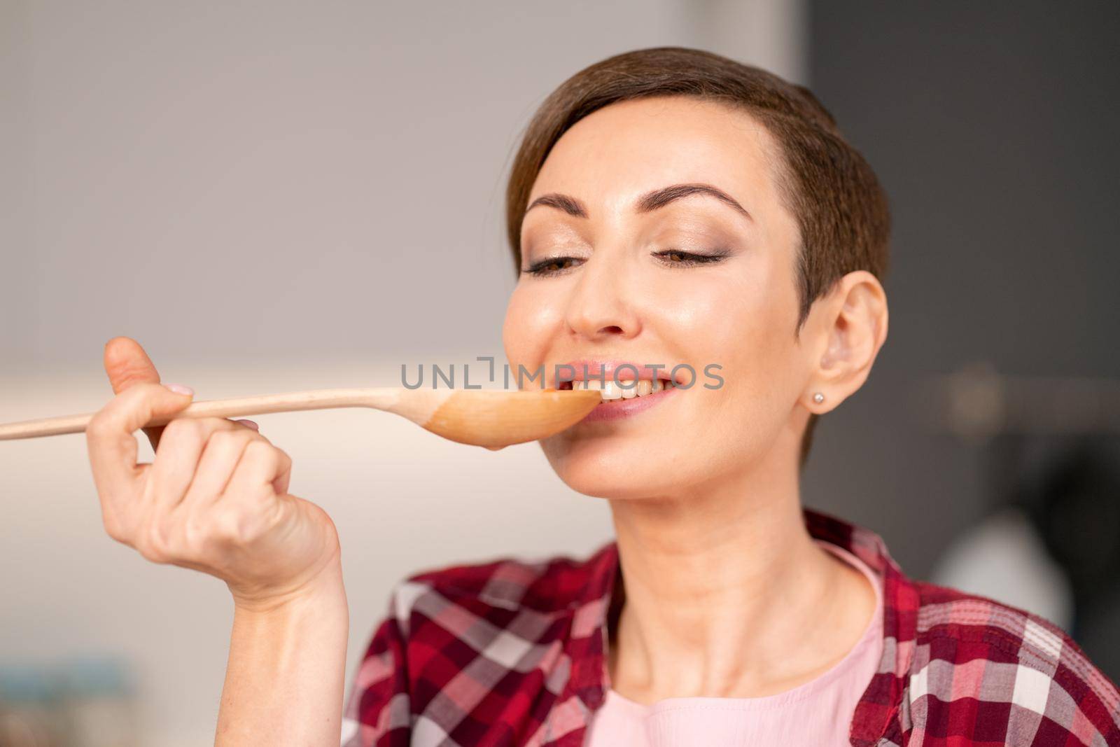 Close-up of a woman trying a cooked dish using a long wooden spoon. Woman with a short hair cooking a dinner for family standing in the modern kitchen of a new house. Healthy food at home. 