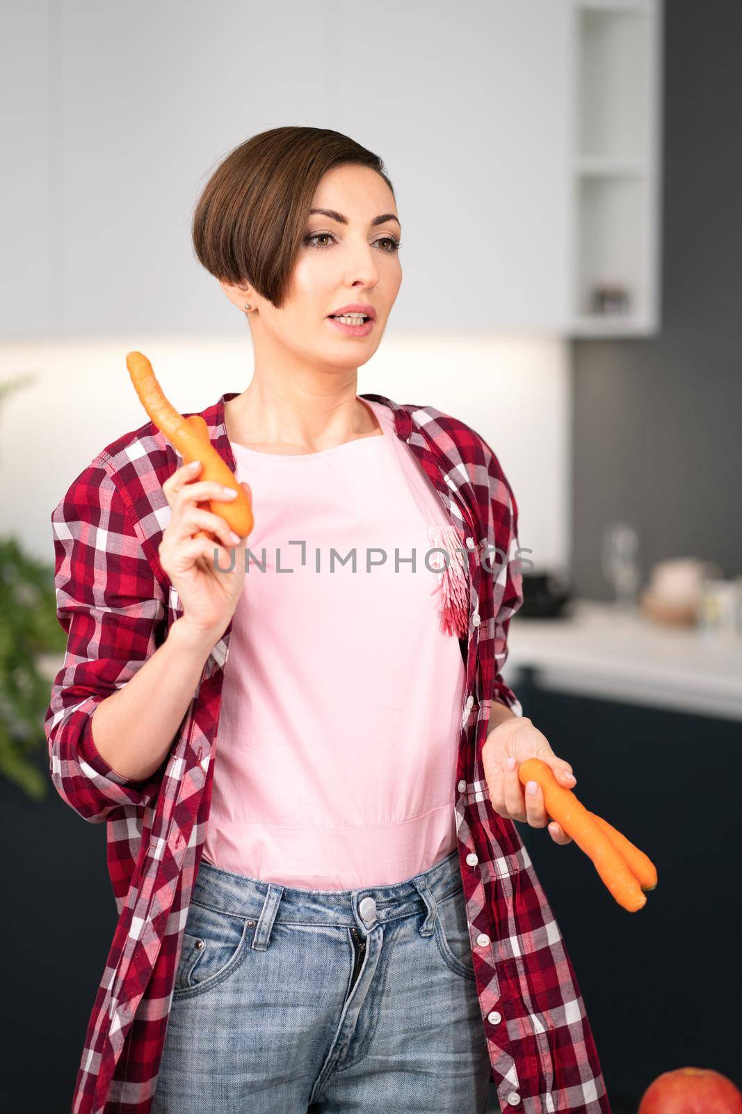 Charming short hair housewife with fresh carrot in the hands prepares carrot cake in the kitchen. Healthy food at home. Healthy food leaving concept by LipikStockMedia